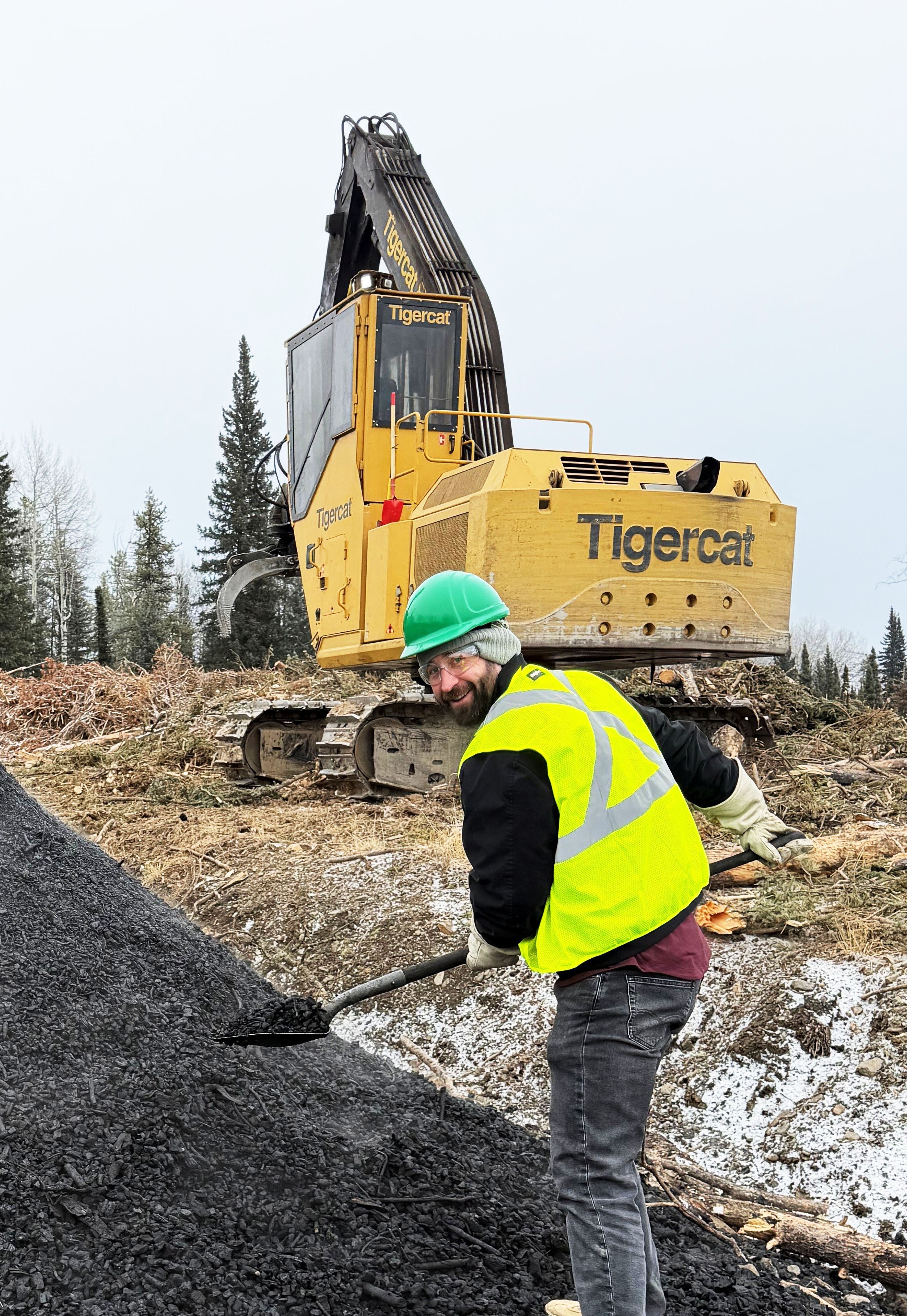 A worker in a yellow safety vest and green hard had shovels carbon near a Tigercat excavator on a work site with trees in the background.