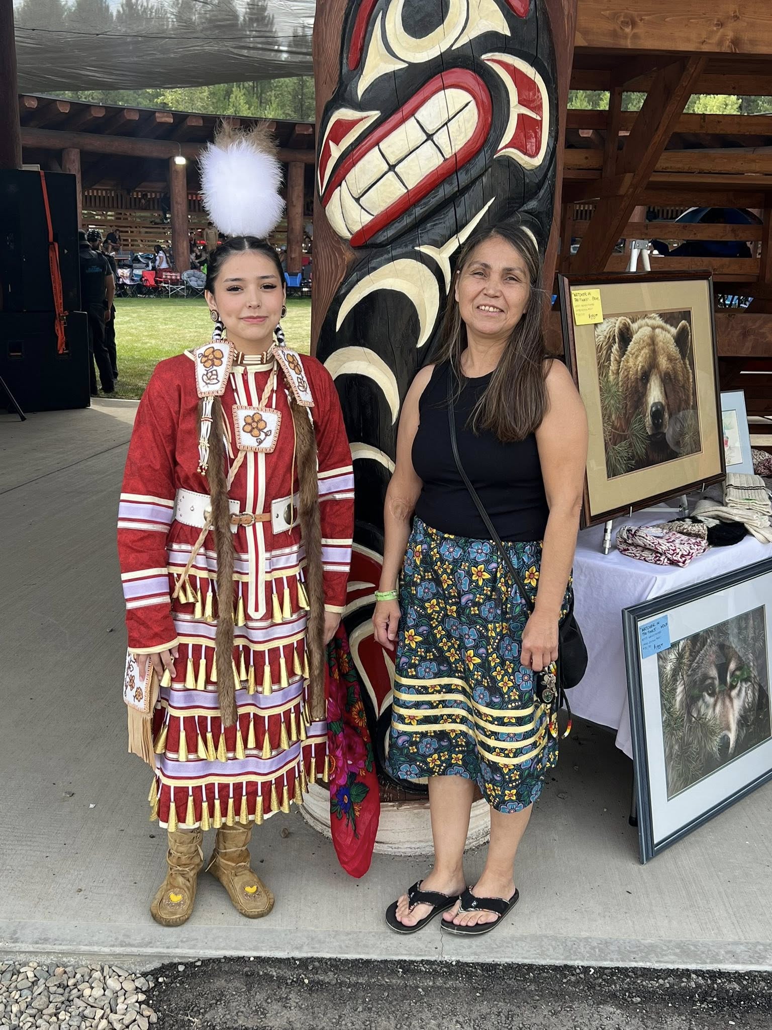 Two women stand side by side; one wears a traditional Indigenous dress, and the other wears a black top and patterned skirt. They are next to a totem pole and art display.