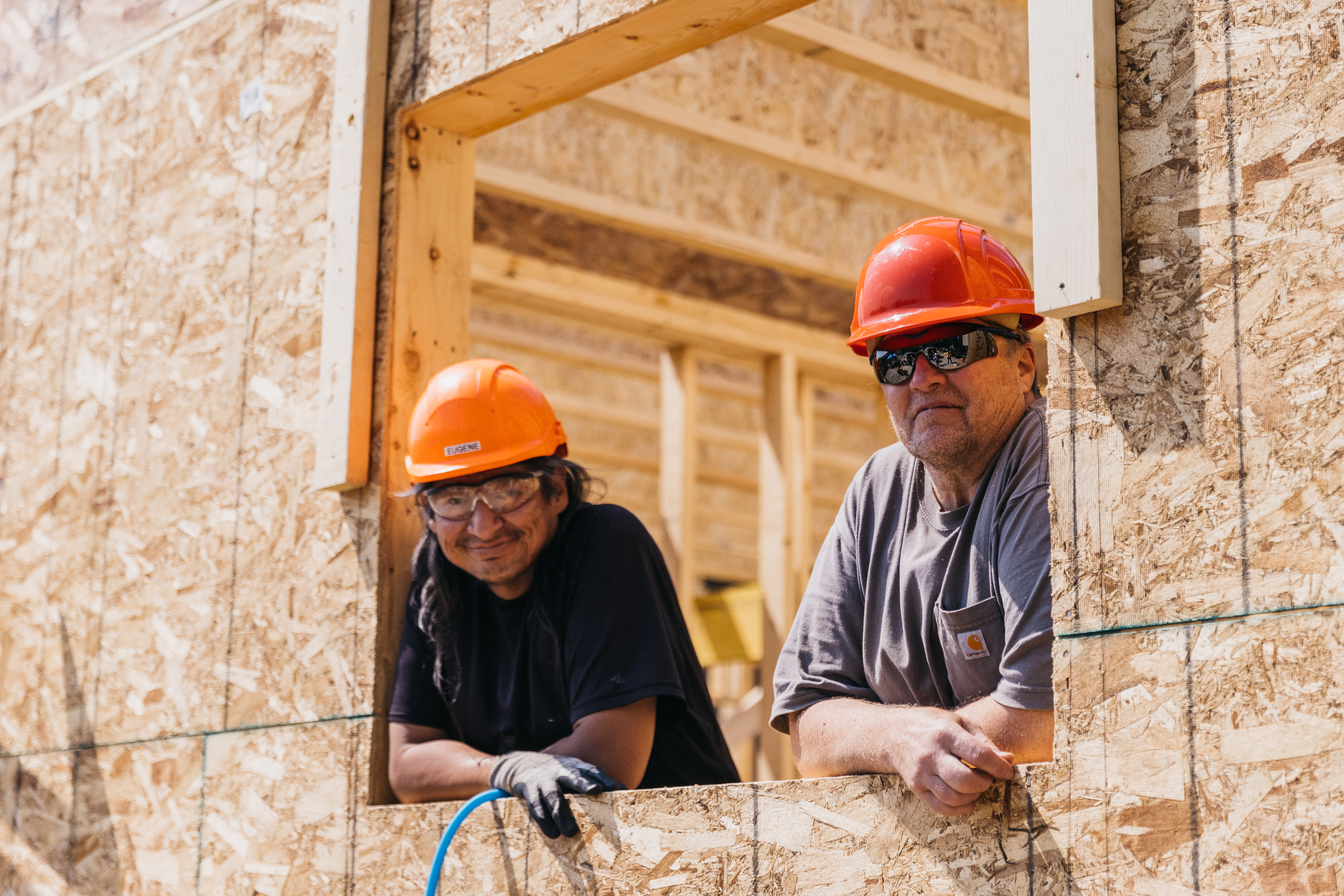 Two construction workers wearing orange hard hats lean out of a framed window in a building under construction.