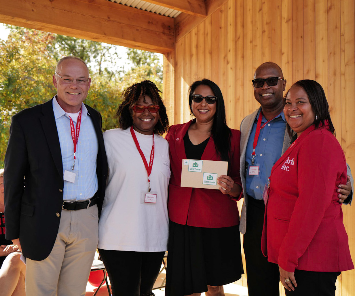 Five people standing together, smiling, under a wooden structure. One person is holding a certificate.