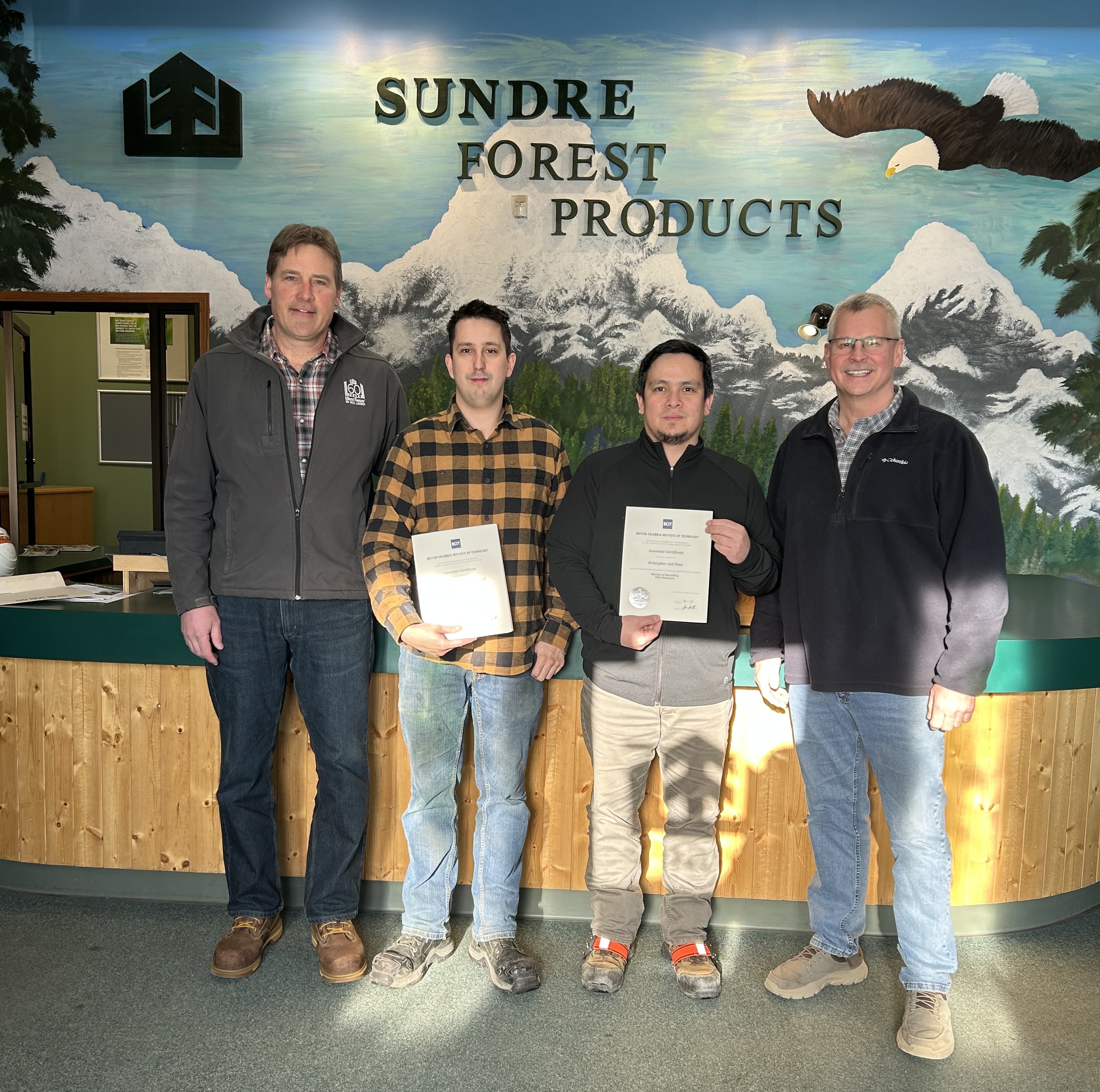Four men stand indoors in front of a Sundre Forest Products sign; two hold certificates, all are facing the camera and smiling.