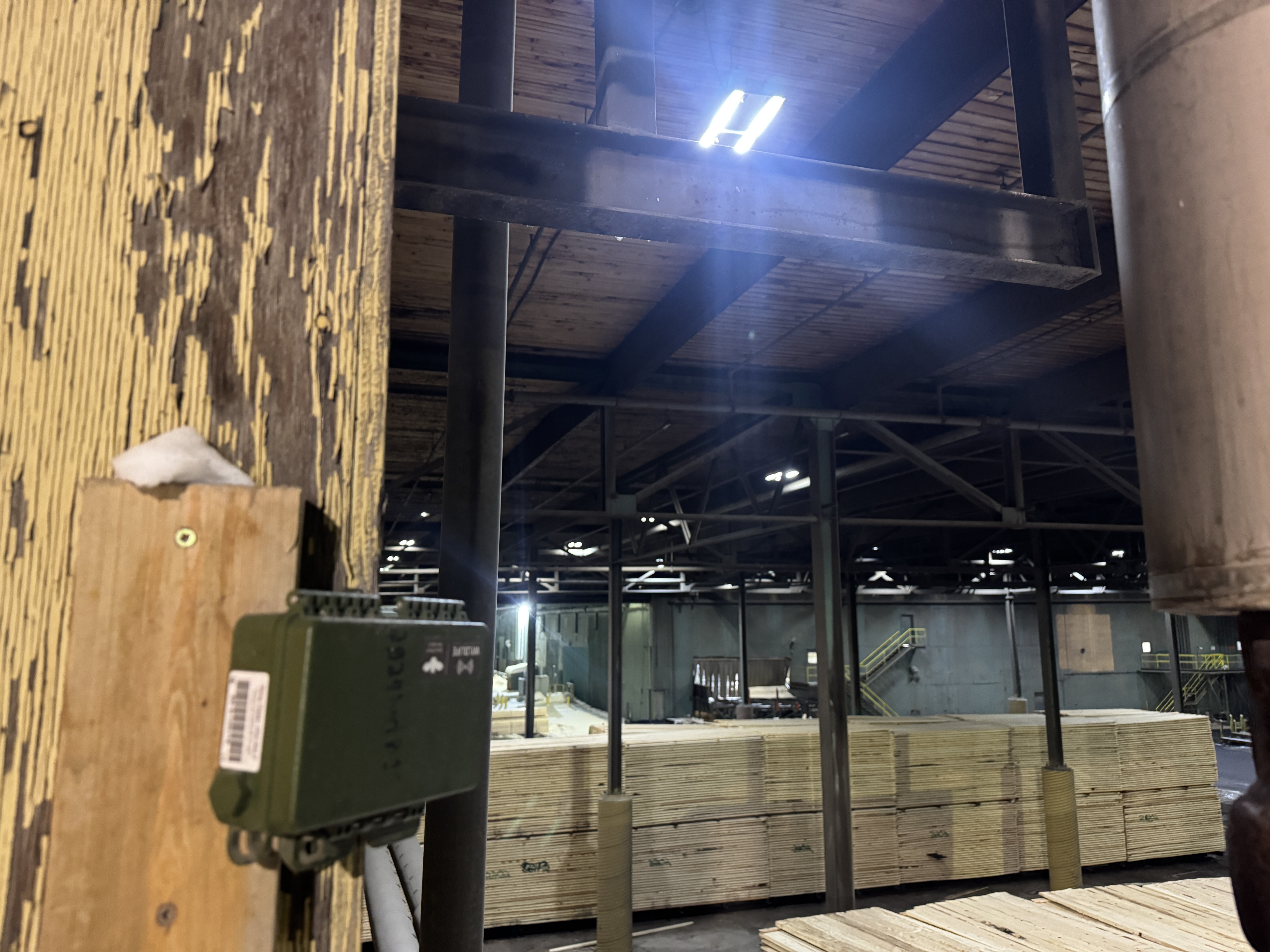 An industrial warehouse interior with stacks of lumber and a green electronic sensor box mounted to a wooden post in the foreground.