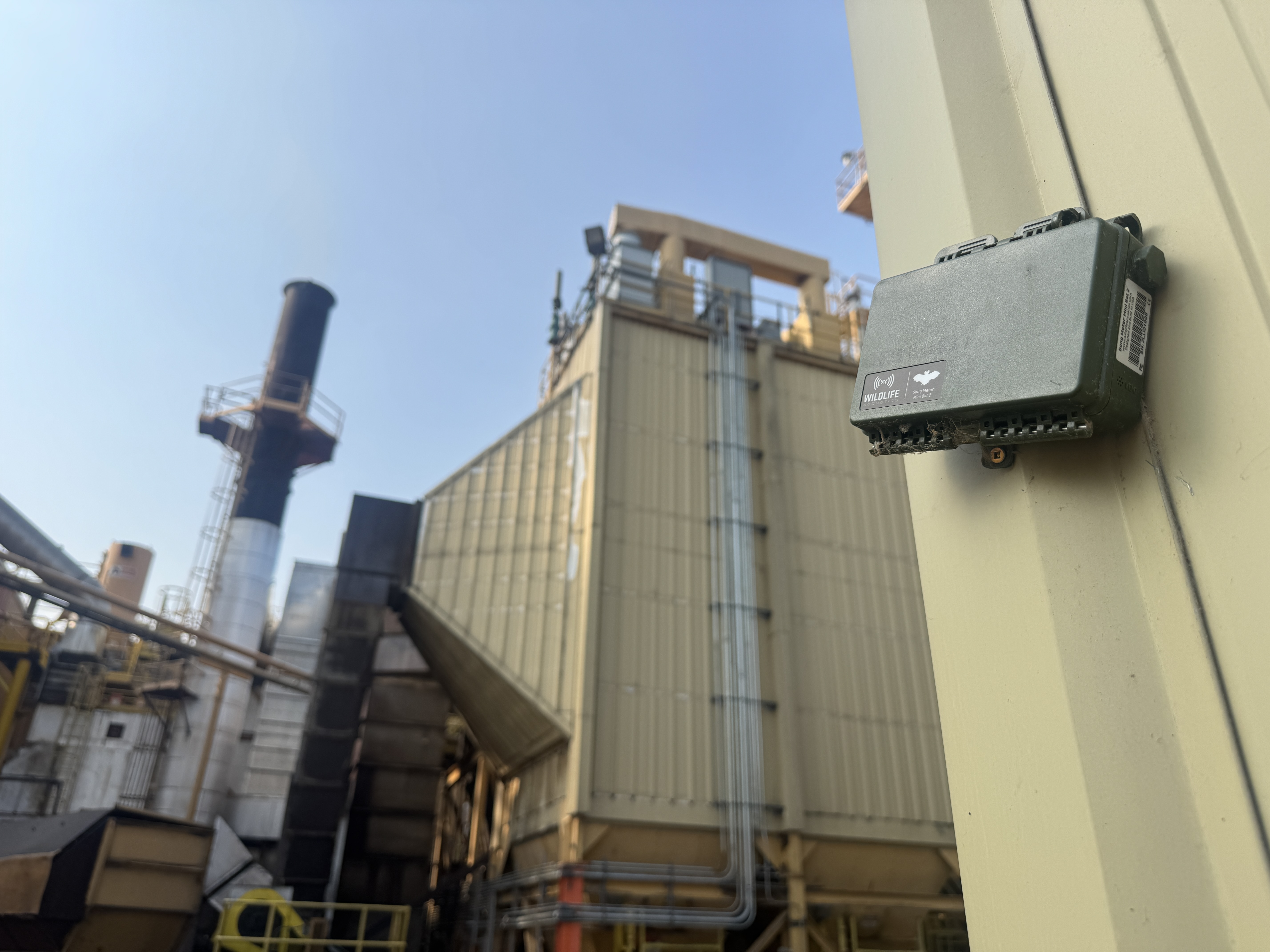 A wildlife acoustic monitoring device mounted on a metal industrial structure with a factory building in the background.