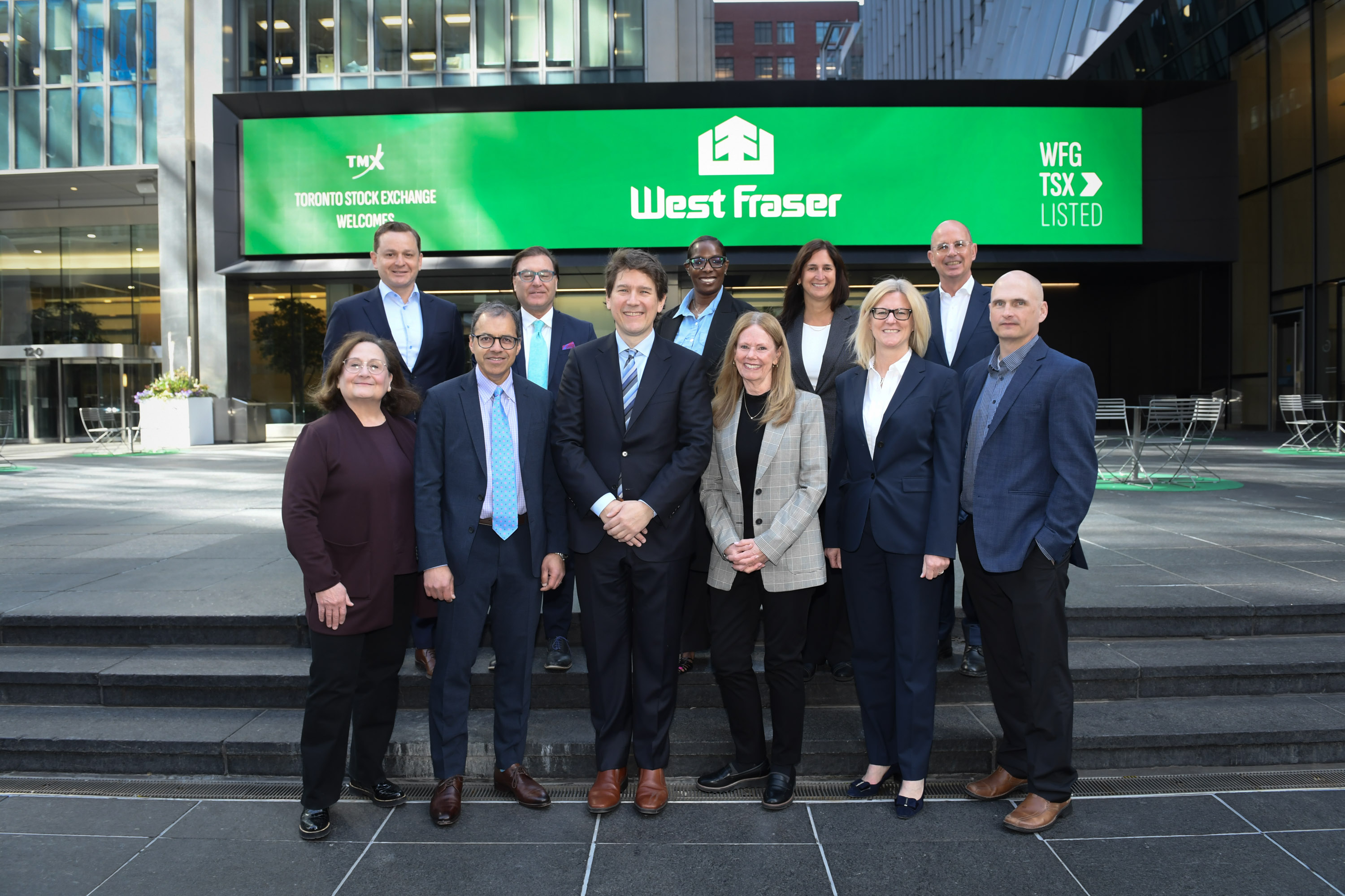 A group of eleven West Fraser team members in business attire stand in front of the Toronto Stock Exchange building with a large green West Fraser sign and the Toronto Stock Exchange logo.