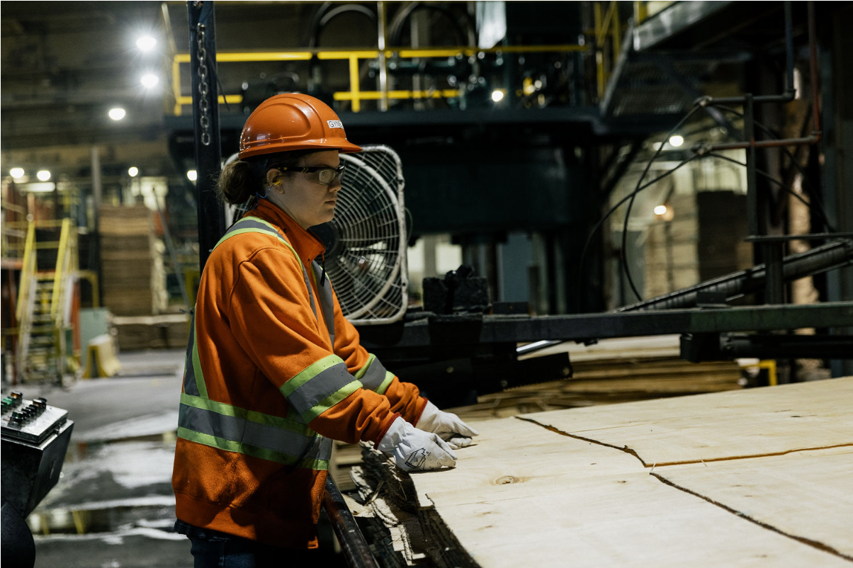 A worker in an orange safety jacket and helmet operates machinery, handling large wooden boards in an industrial setting.