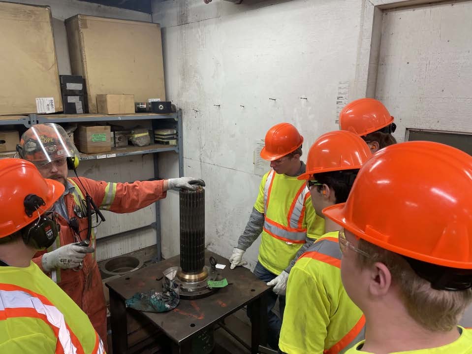 Four students and one worker in reflective orange and yellow safety vests and orange hard hats stand in a circle in an industrial mill setting, while the worker teaches the students.