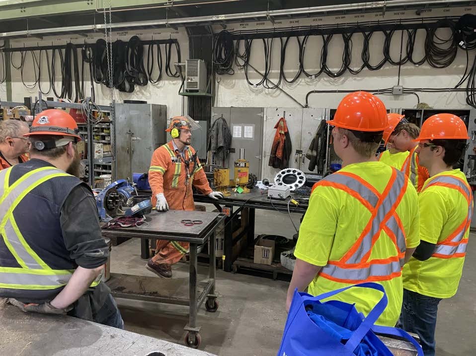 A group of students and mill workers, all wearing hard hats and safety vests, tour an industrial mill setting and workshop area.