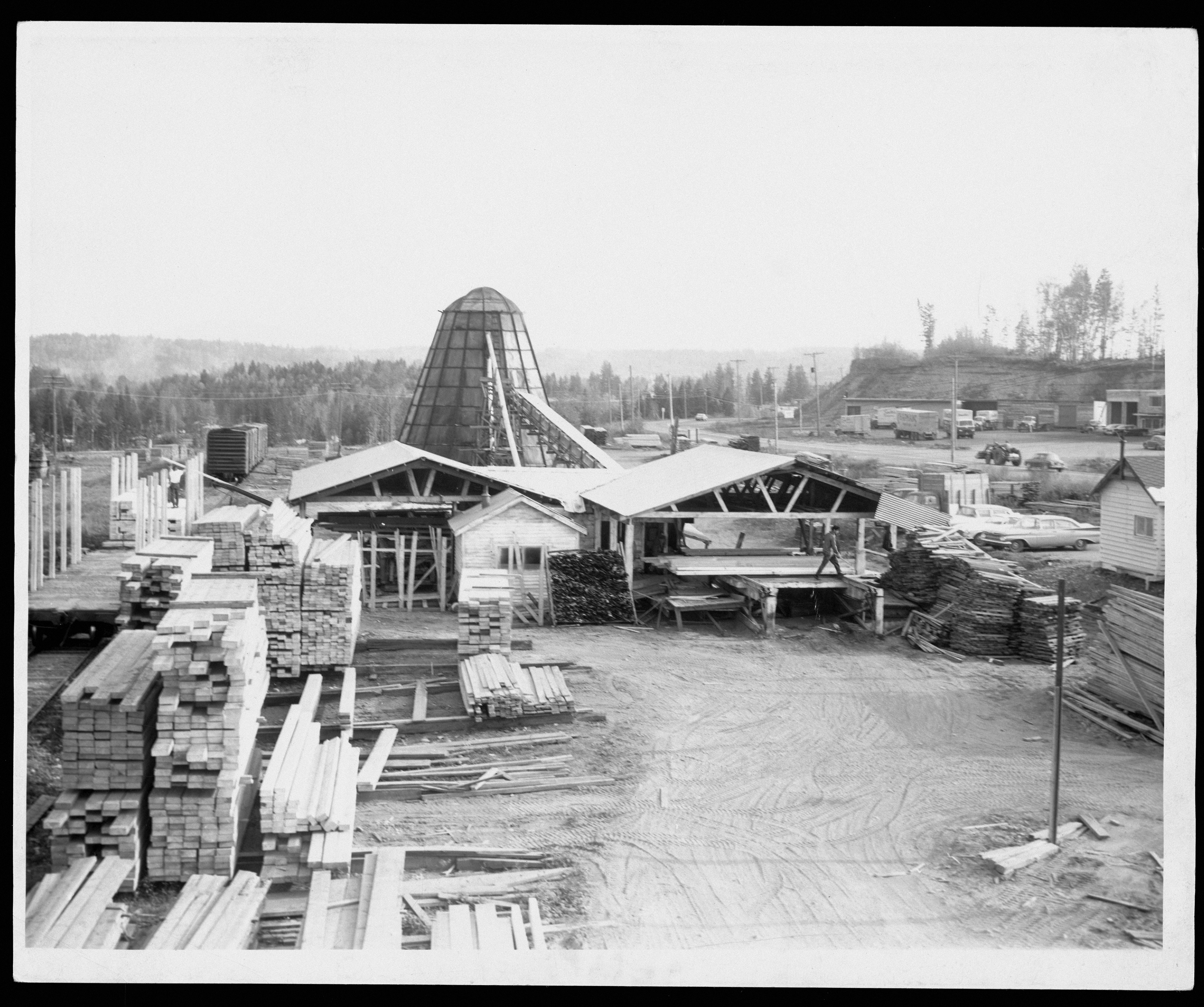 Black and white photo of a lumber yard with stacked wood, buildings, and a large conical structure in the center; trees and vehicles are visible in the background.