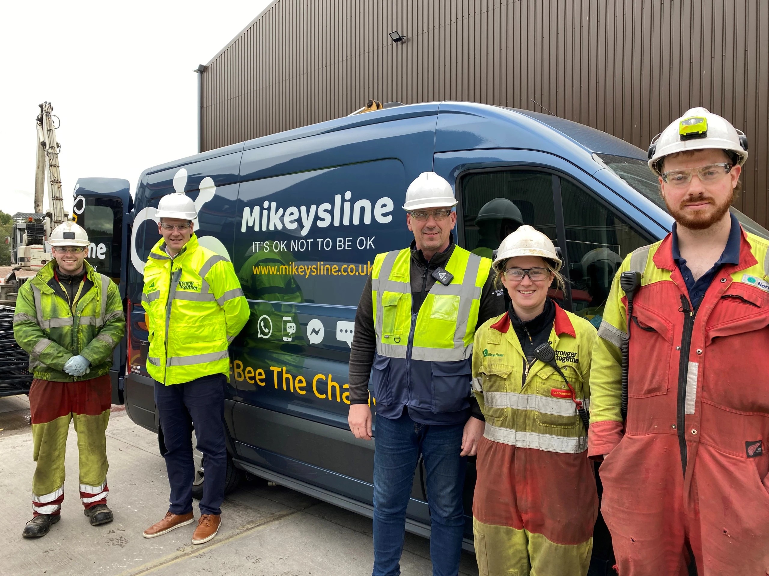 Five people in safety gear stand in front of a van with “Mikeysline” branding and mental health awareness messages.
