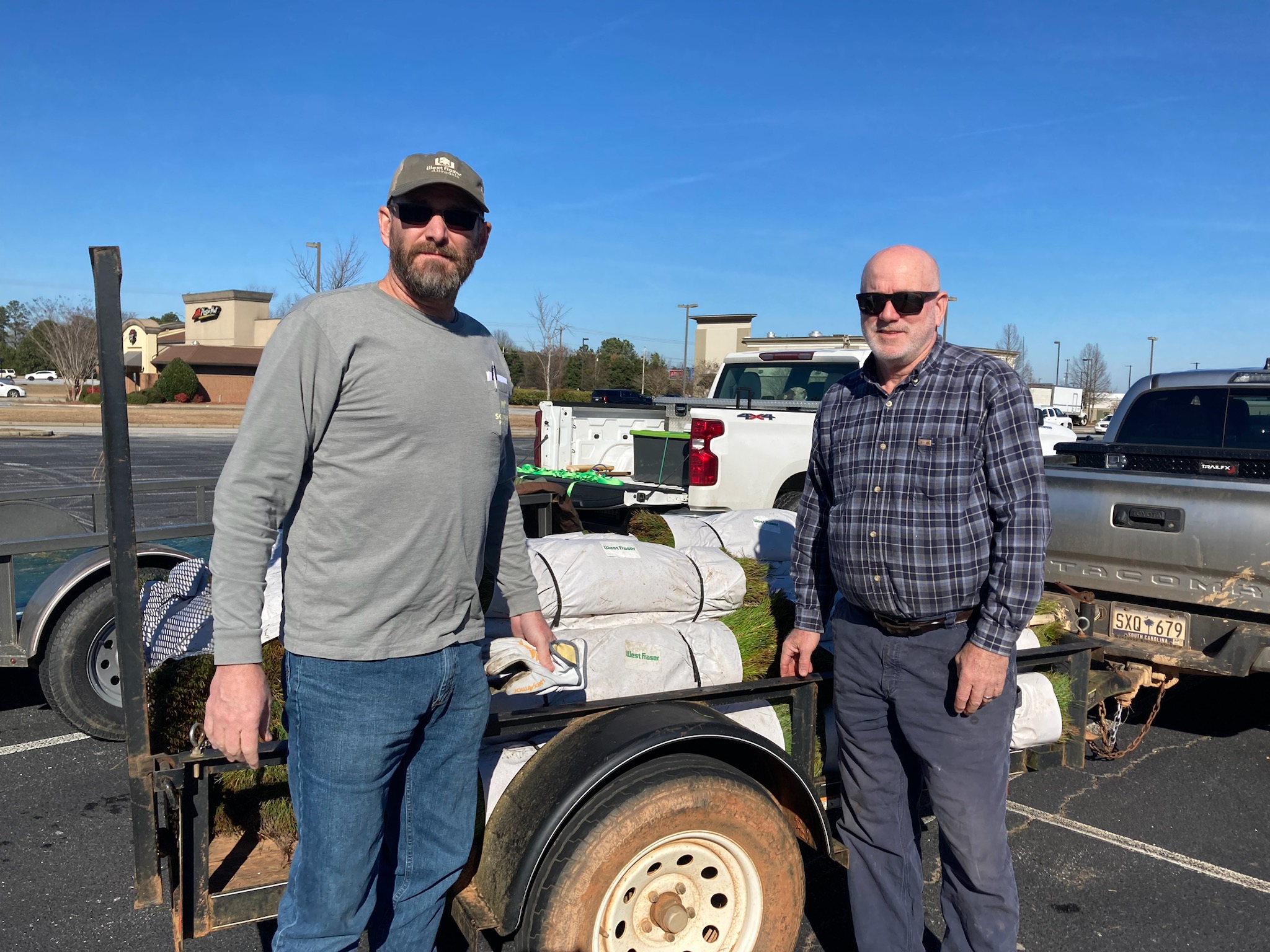 Two men stand by a trailer loaded with bags and sod in a parking lot on a sunny day.