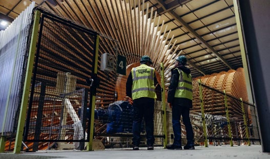 Two workers in yellow safety vests and green hard hats stand in a mill, in front of a gate, looking at wood samples.