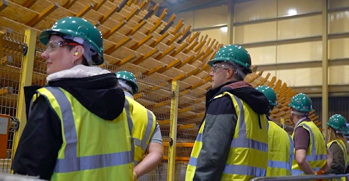 A group of people wearing safety vests, hard hats, and ear protection tour an industrial facility with stacks of wood in the background.