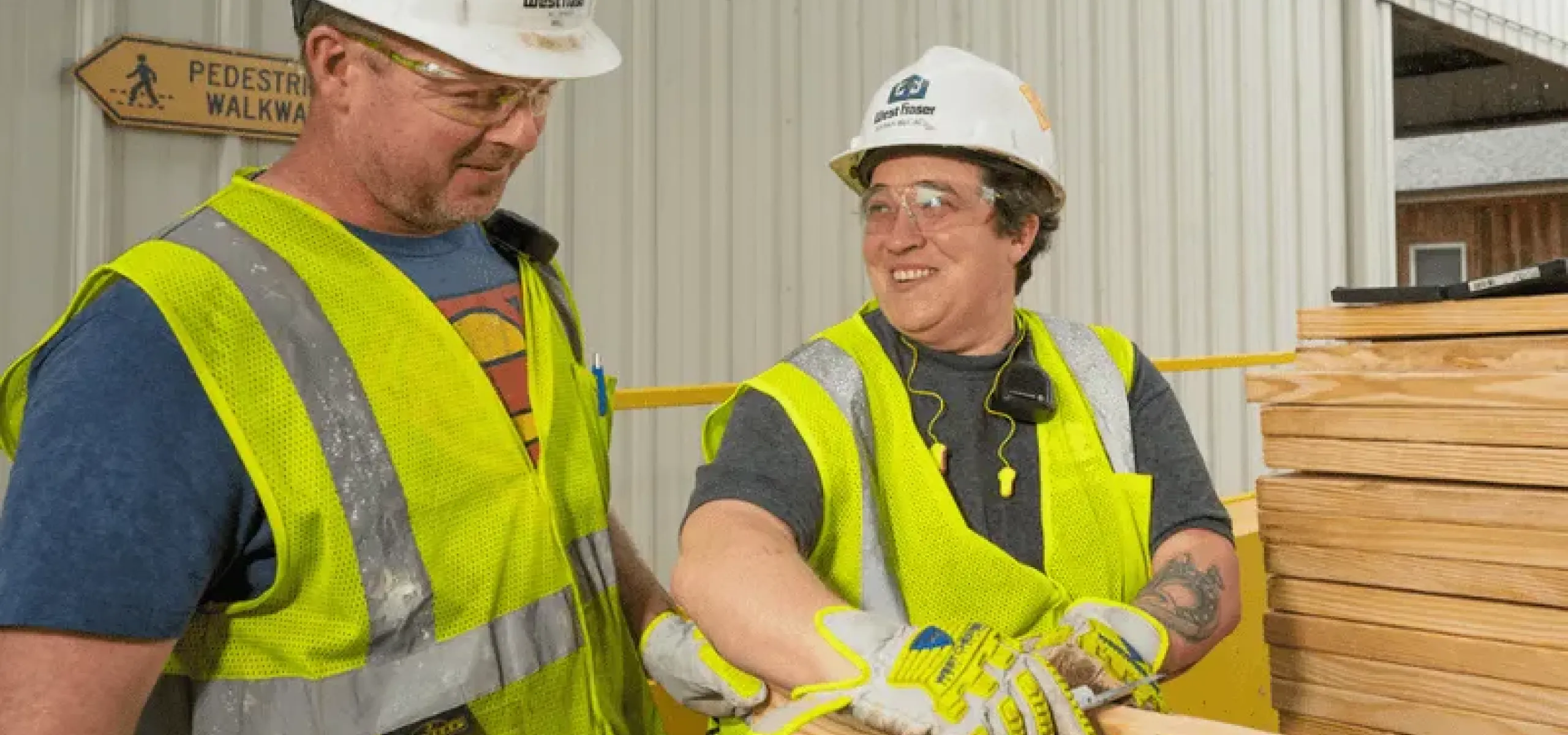 Two construction workers in safety vests, gloves, and helmets are handling wooden planks outside a building. One worker is smiling and talking to the other. A "Pedestrian Walkway" sign is visible.