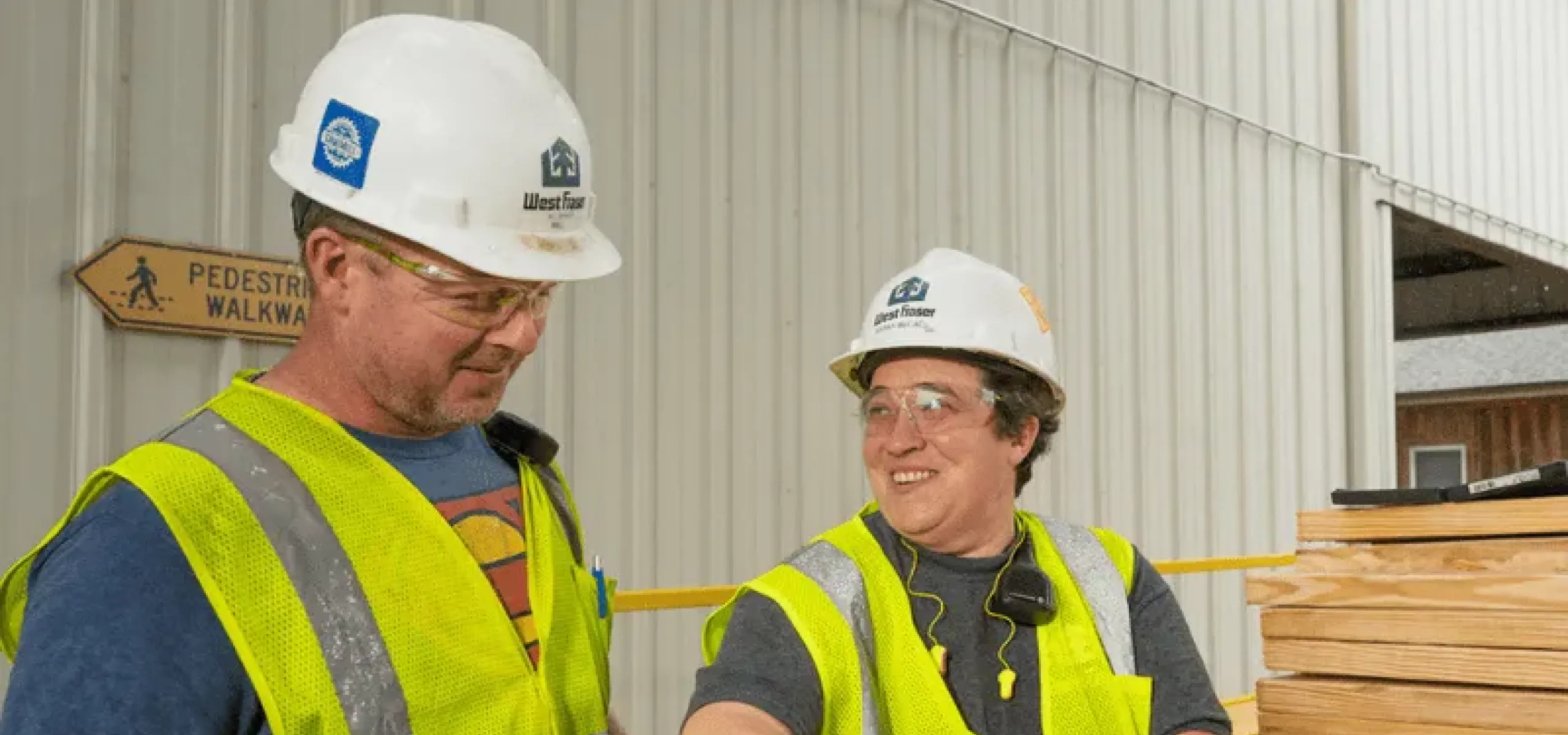 Two construction workers in safety vests, gloves, and helmets are handling wooden planks outside a building. One worker is smiling and talking to the other. A "Pedestrian Walkway" sign is visible.
