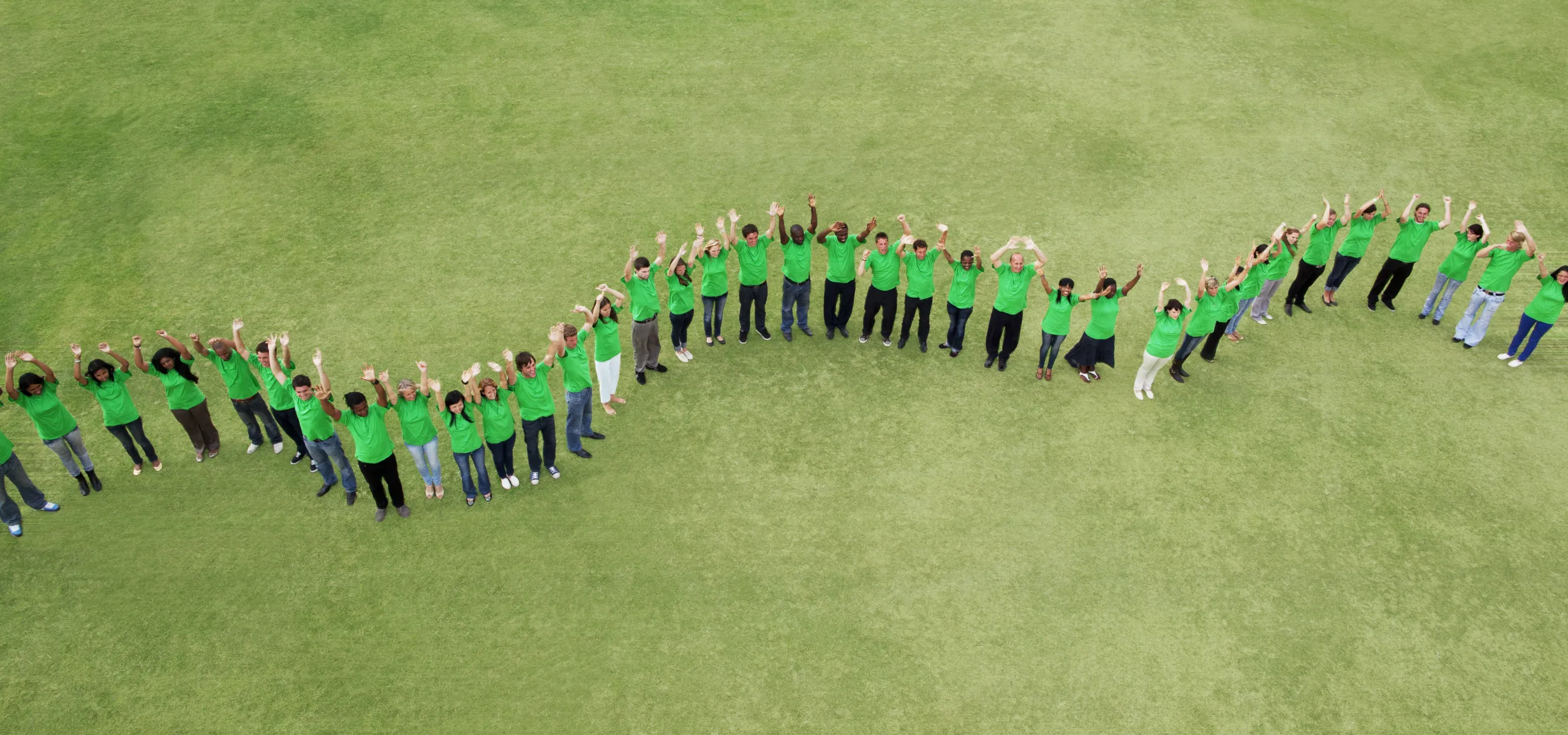 A group of people wearing green shirts stand outside on grass, forming a wavy line and raising their hands in the air.
