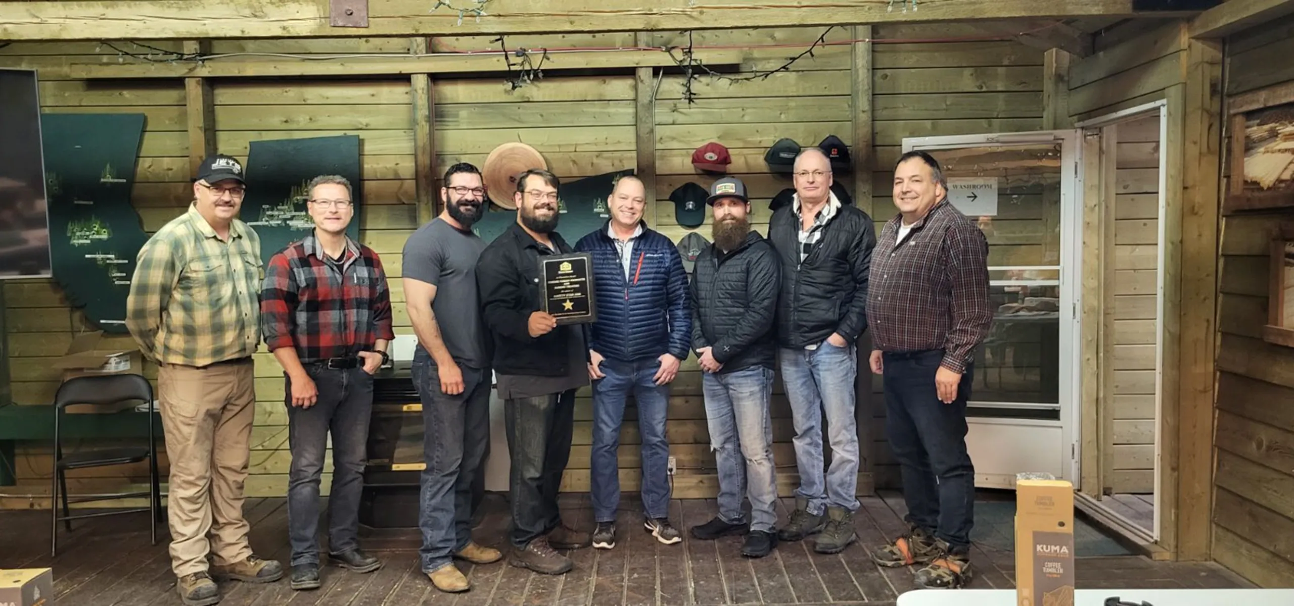 A group of eight men standing indoors, one holding a plaque. They are in a rustic wooden room with hats displayed on the wall and boxes on the floor.