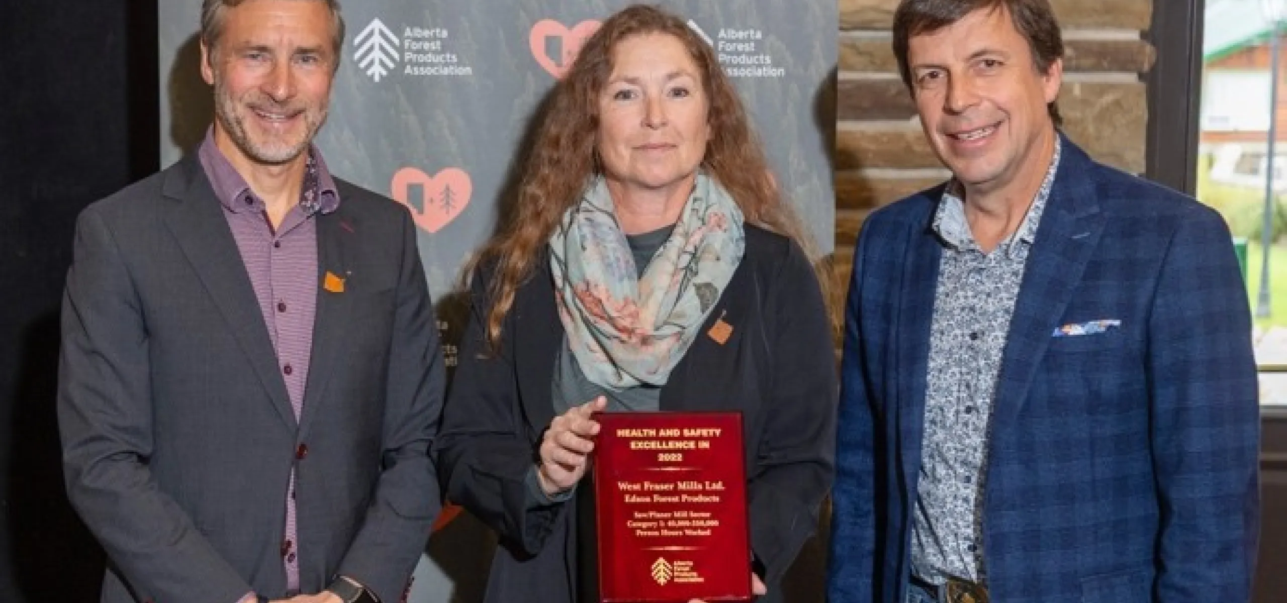 Three individuals pose together, with the middle person holding a red award plaque. They stand in front of a backdrop with logos from the Alberta Forest Products Association.