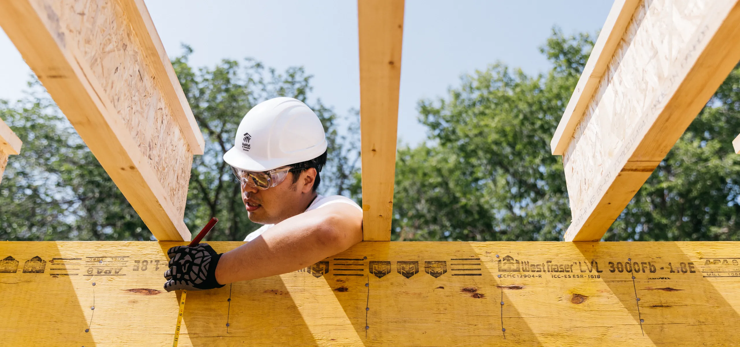 A construction worker wearing a white hard hat and safety glasses measures and marks a wooden beam at a construction site.