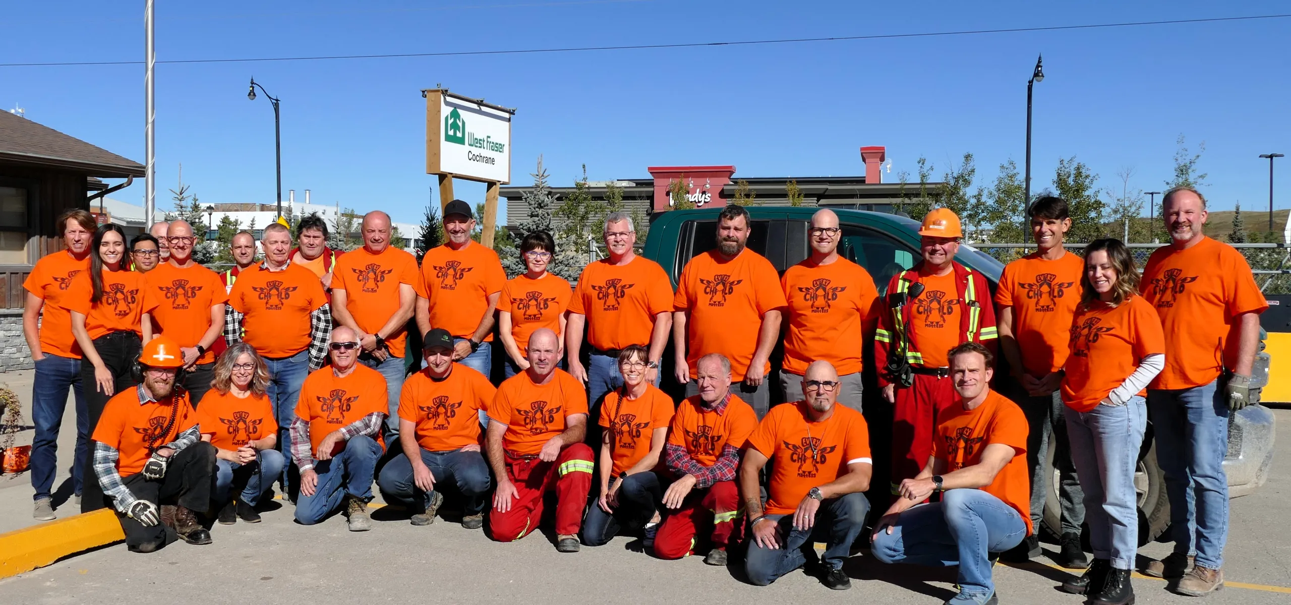 A group of people wearing orange shirts and outdoor gear pose for a photo in front of a building under a clear blue sky.