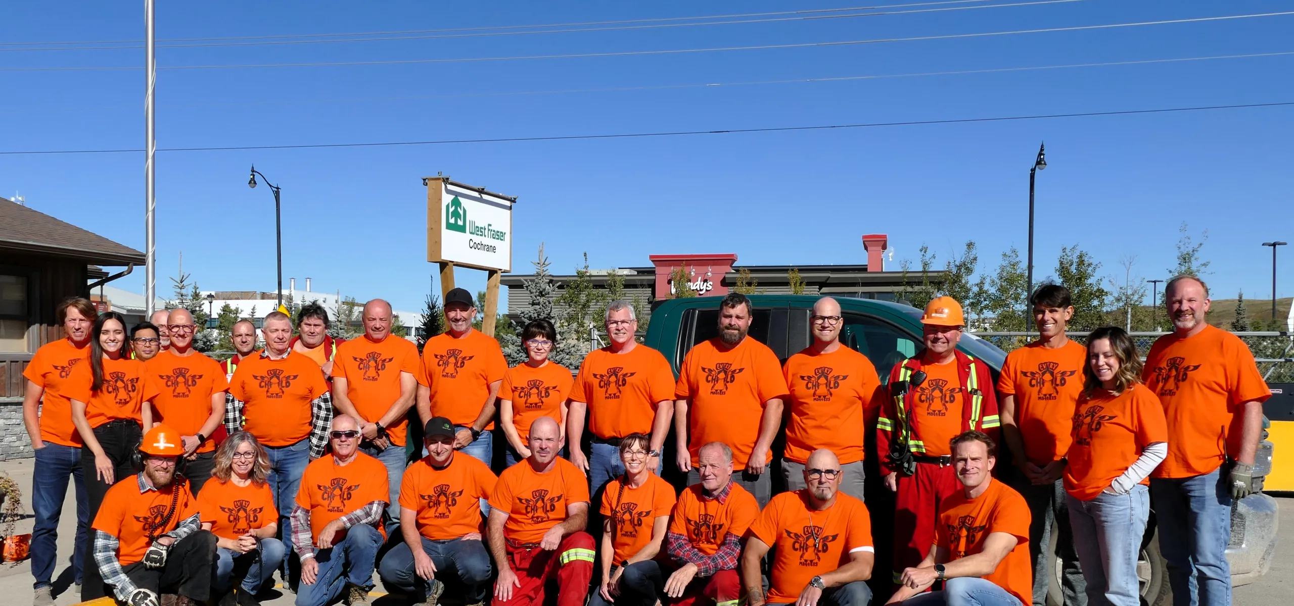 A group of people wearing orange shirts and outdoor gear pose for a photo in front of a building under a clear blue sky.