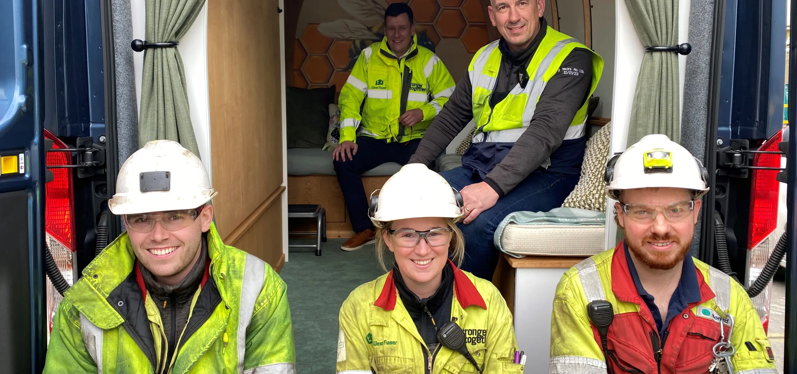 Four workers in safety gear smile for a group photo, seated in the back of a van with a wooden interior. Two additional people sit behind them.