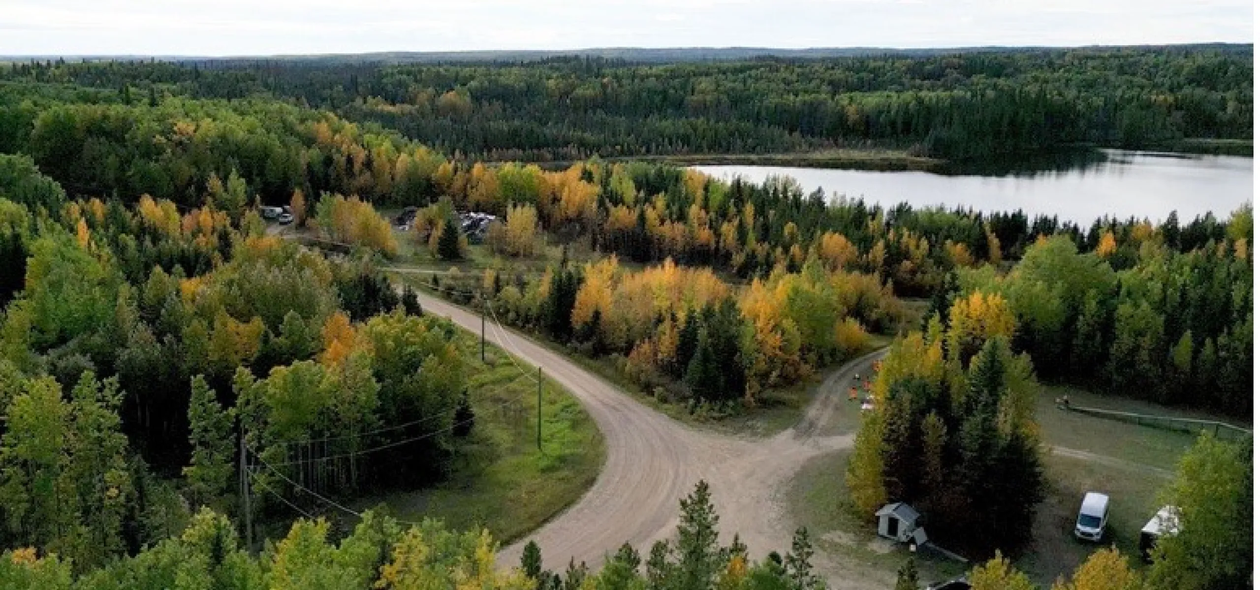 Aerial view of a forest landscape with a dirt road intersection, surrounded by trees with autumn foliage, and a lake visible in the background.