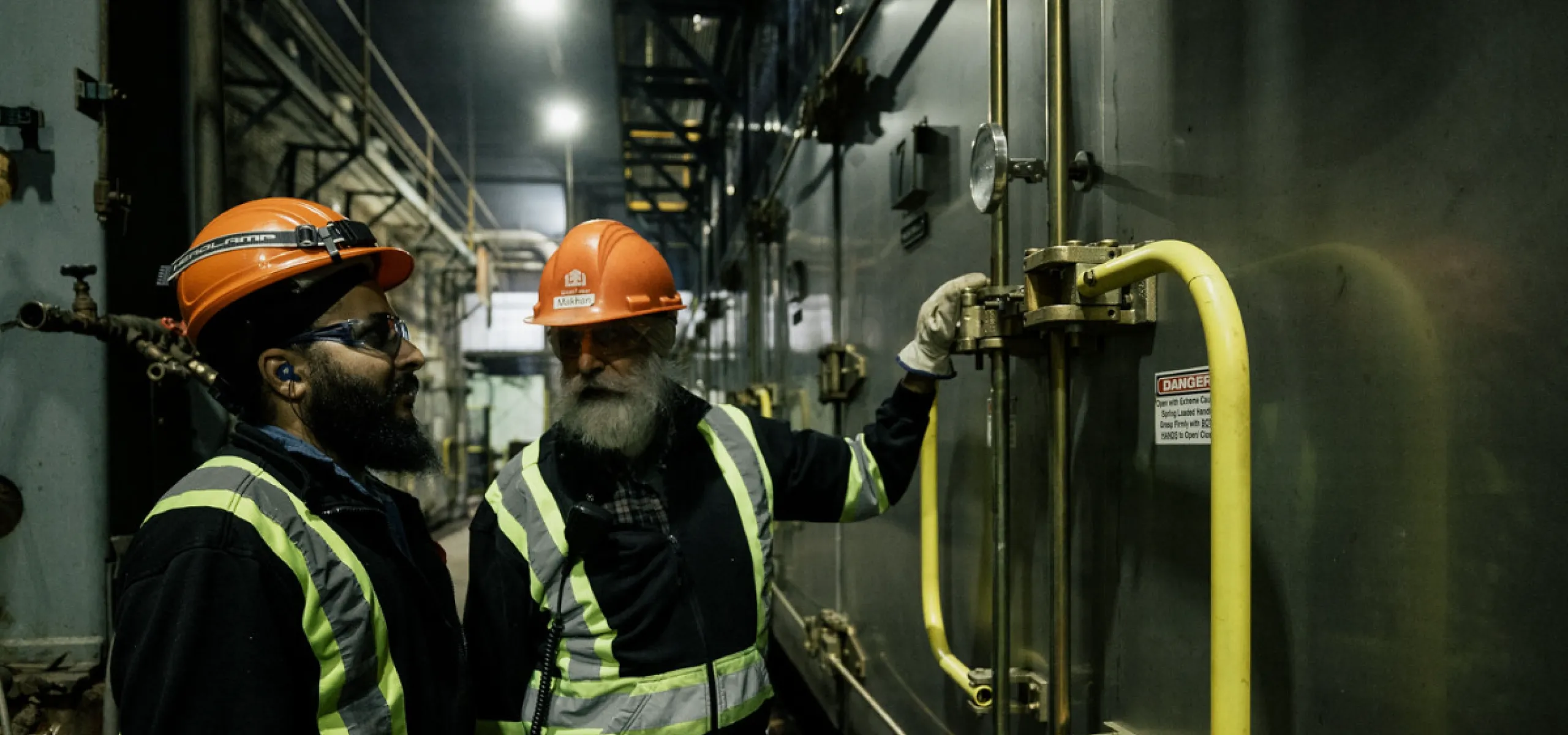 Two workers in safety gear inspect a large industrial machine inside a dimly lit facility.