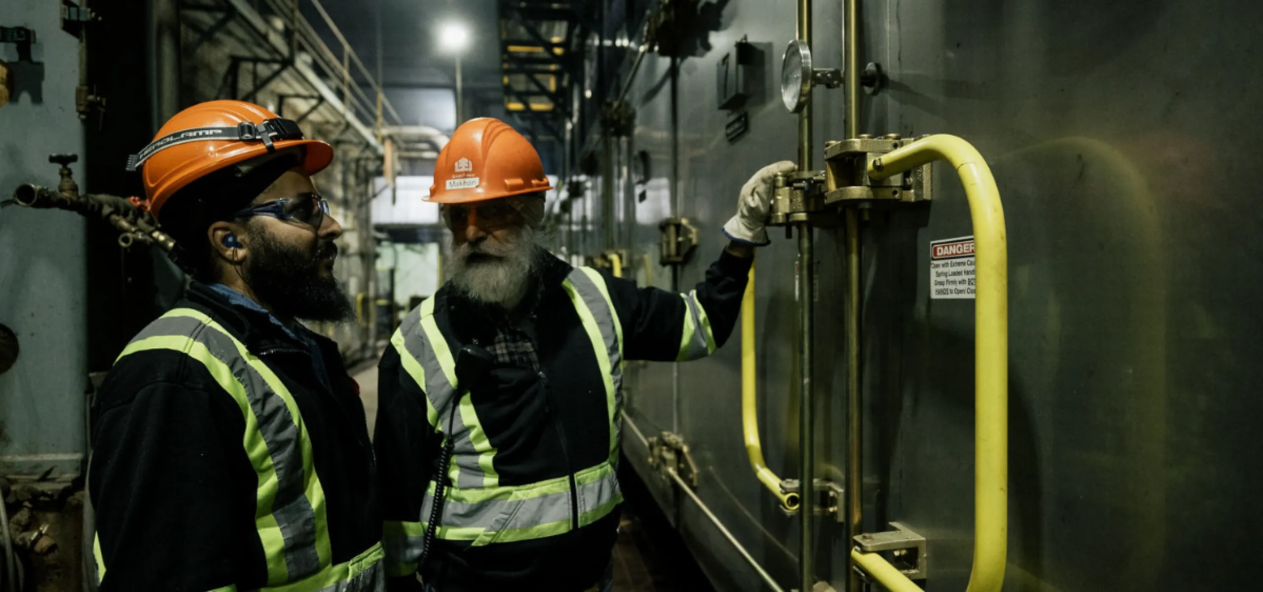 Two workers in safety gear inspect a large industrial machine inside a dimly lit facility.