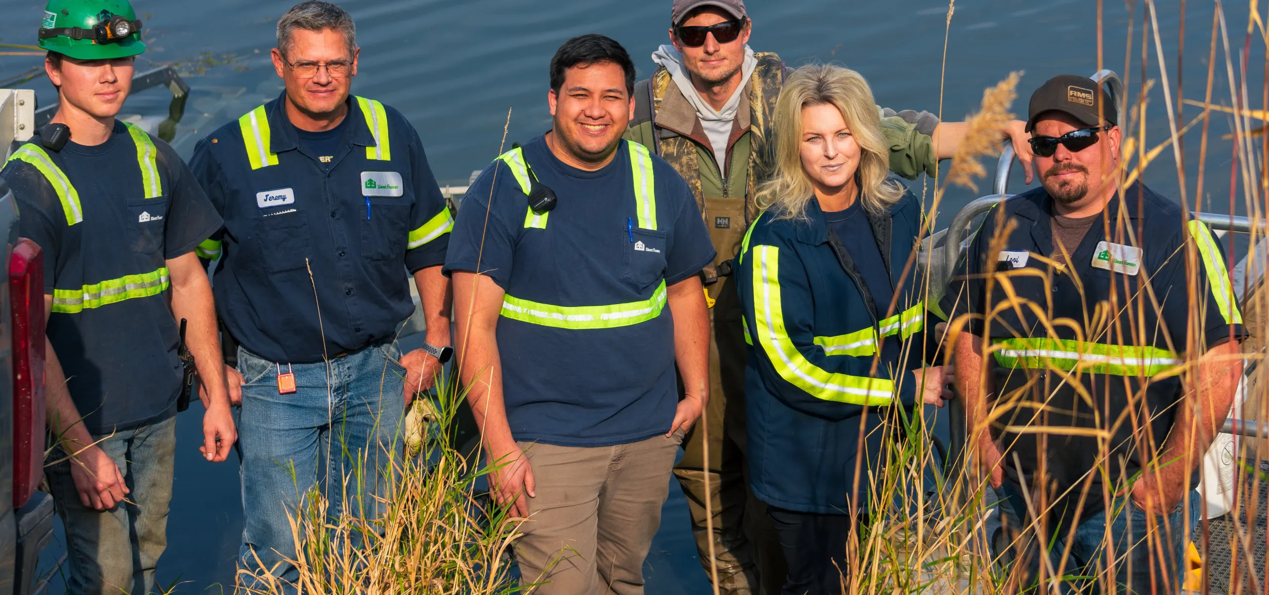 A group of six people wearing safety vests and work attire stand near water with tall grass in the foreground.
