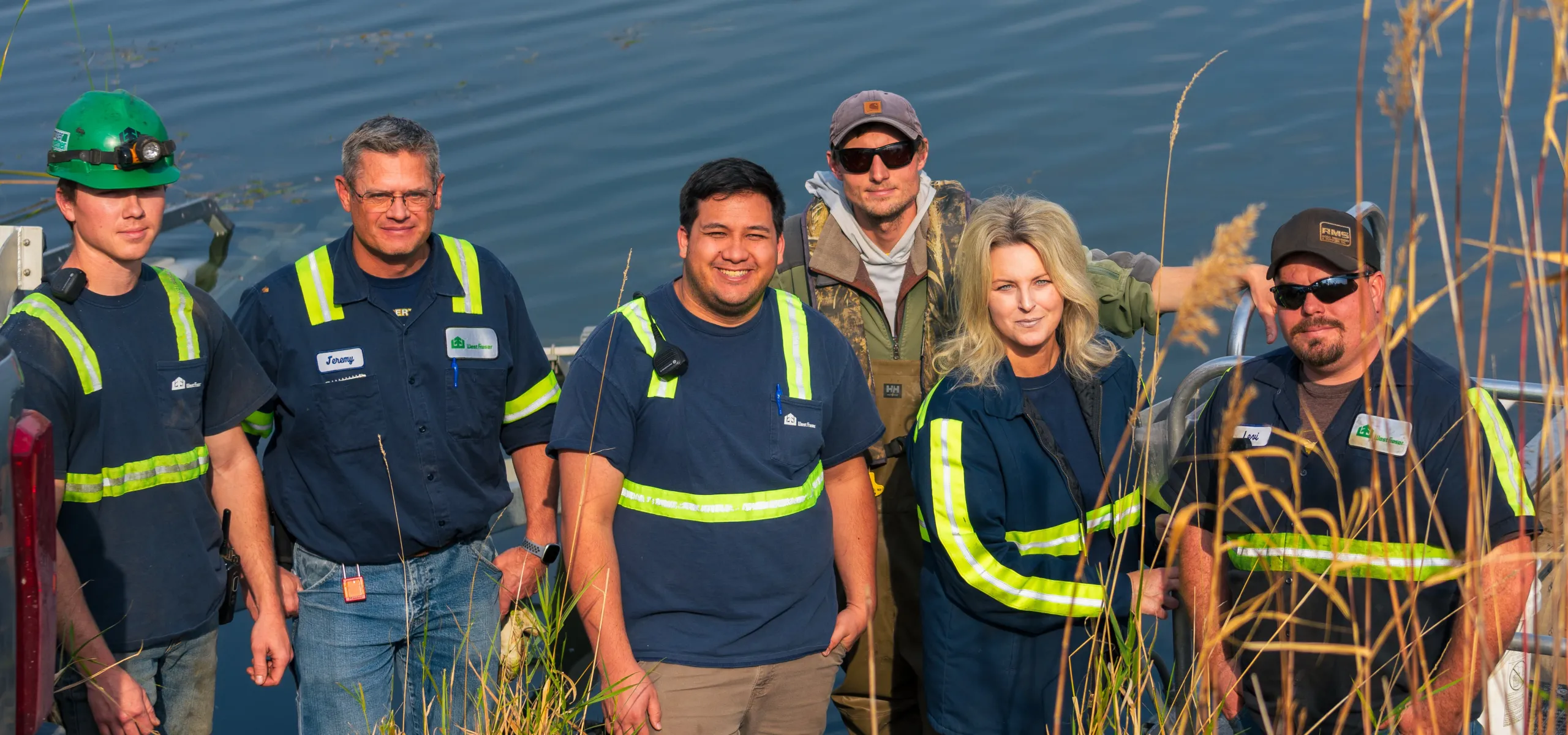 A group of six people wearing safety vests and work attire stand near water with tall grass in the foreground.