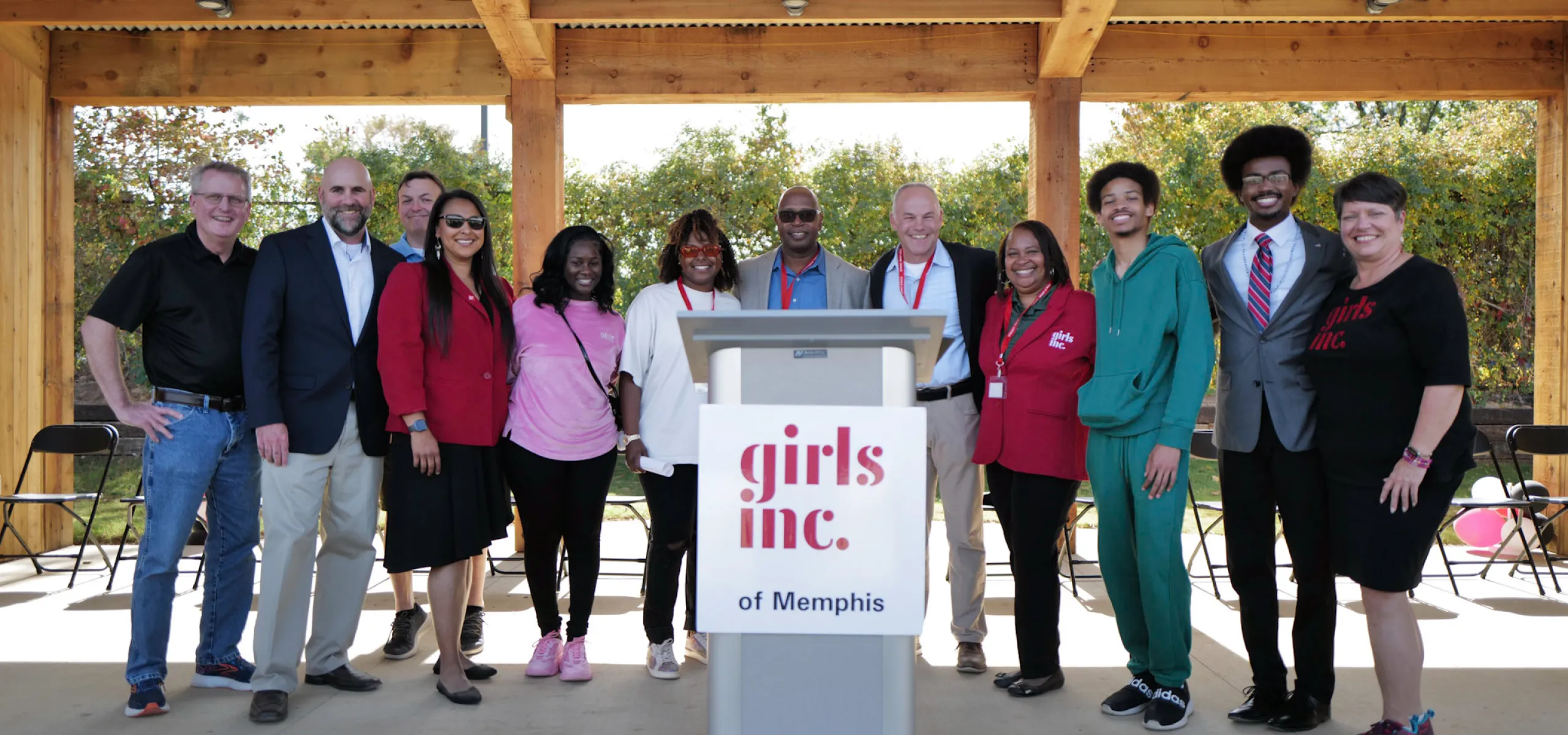 A group of people stands together under a wooden pavilion, with a podium labeled "Girls Inc. of Memphis" in front.