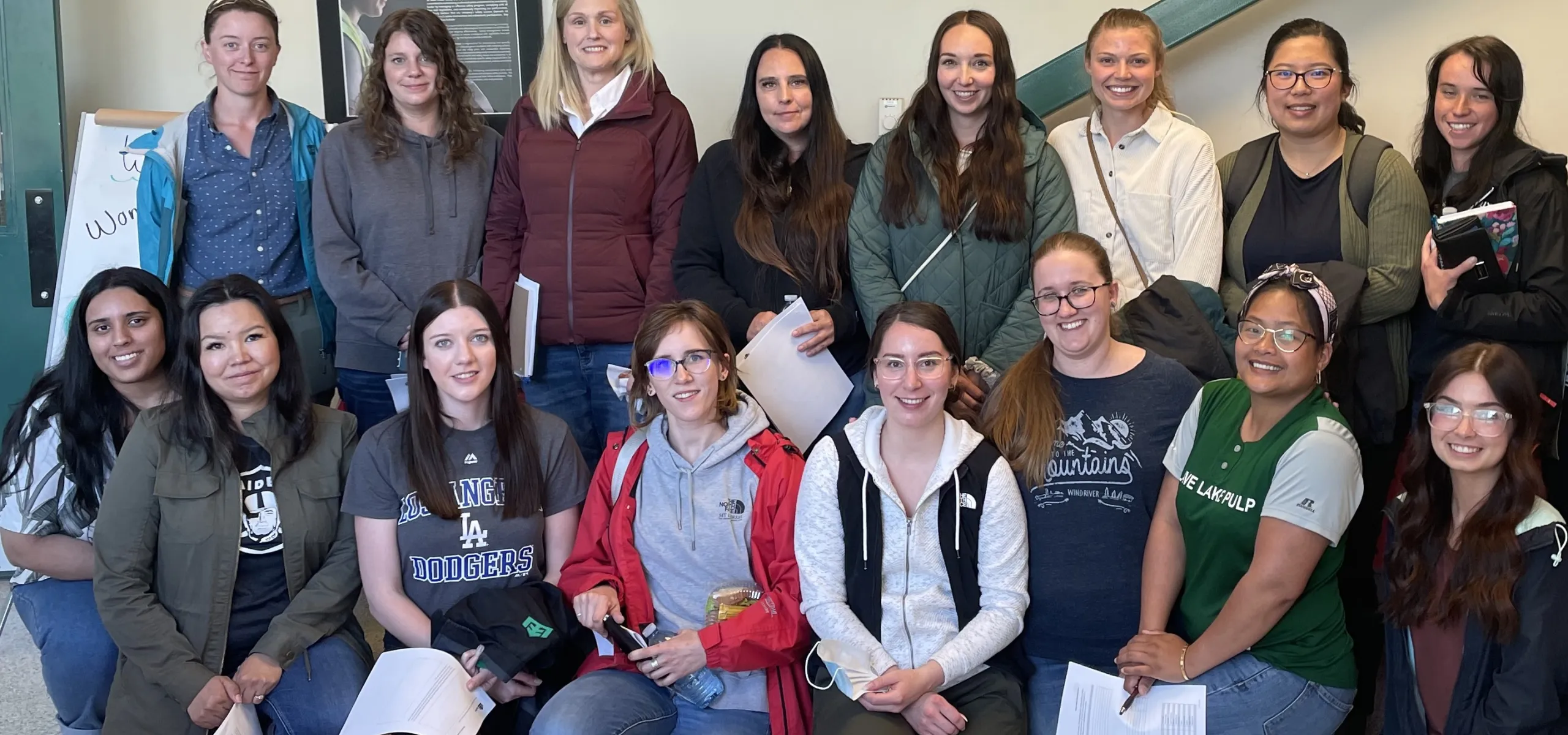 Group of people posing indoors, some holding papers. They stand and kneel in two rows. A staircase and a wall are in the background.