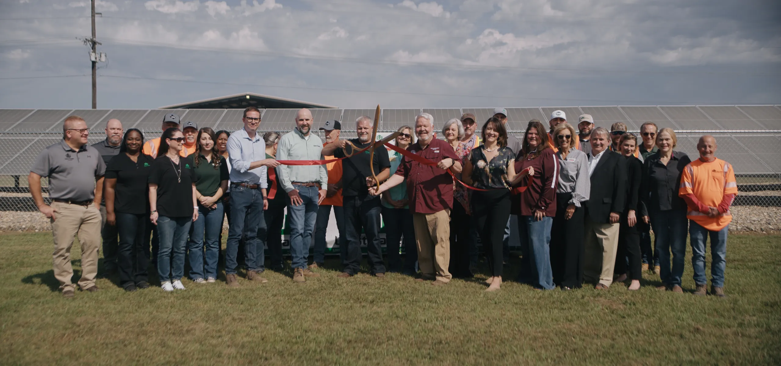 A group of people stand together outside in front of solar panels. One person is cutting a ribbon with large scissors. It is a sunny day with clouds in the sky.