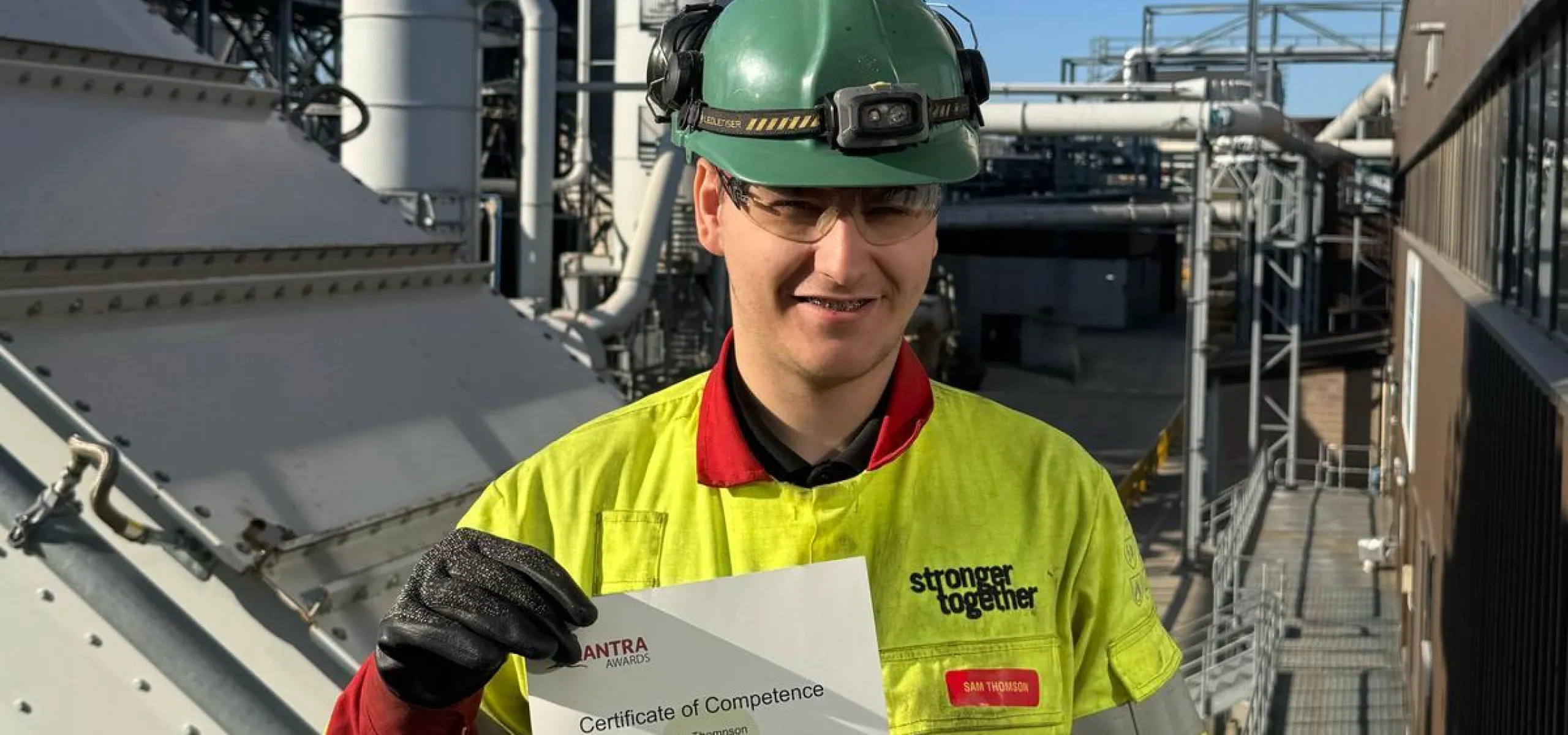 A worker wearing safety gear and holding a certificate stands in front of industrial equipment with smoke rising from a stack.