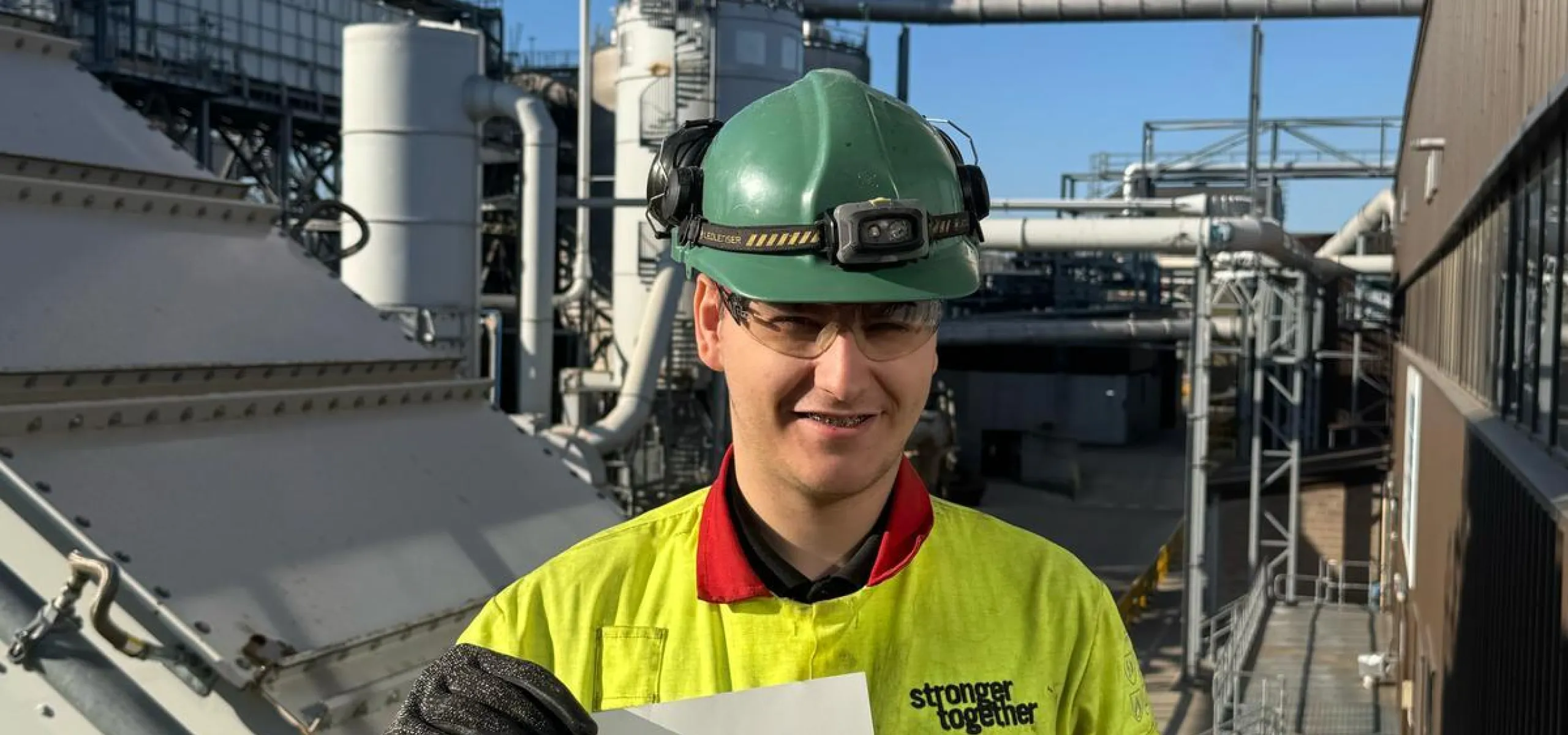 A worker wearing safety gear and holding a certificate stands in front of industrial equipment with smoke rising from a stack.