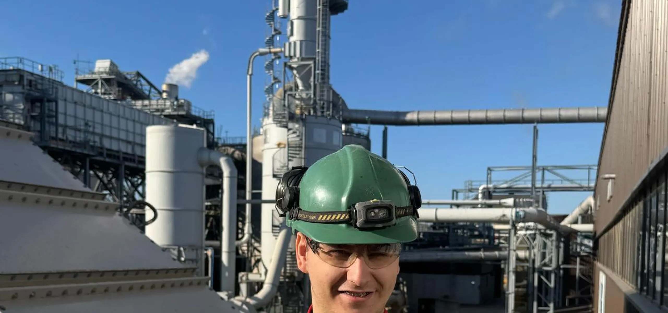 A worker wearing safety gear and holding a certificate stands in front of industrial equipment with smoke rising from a stack.