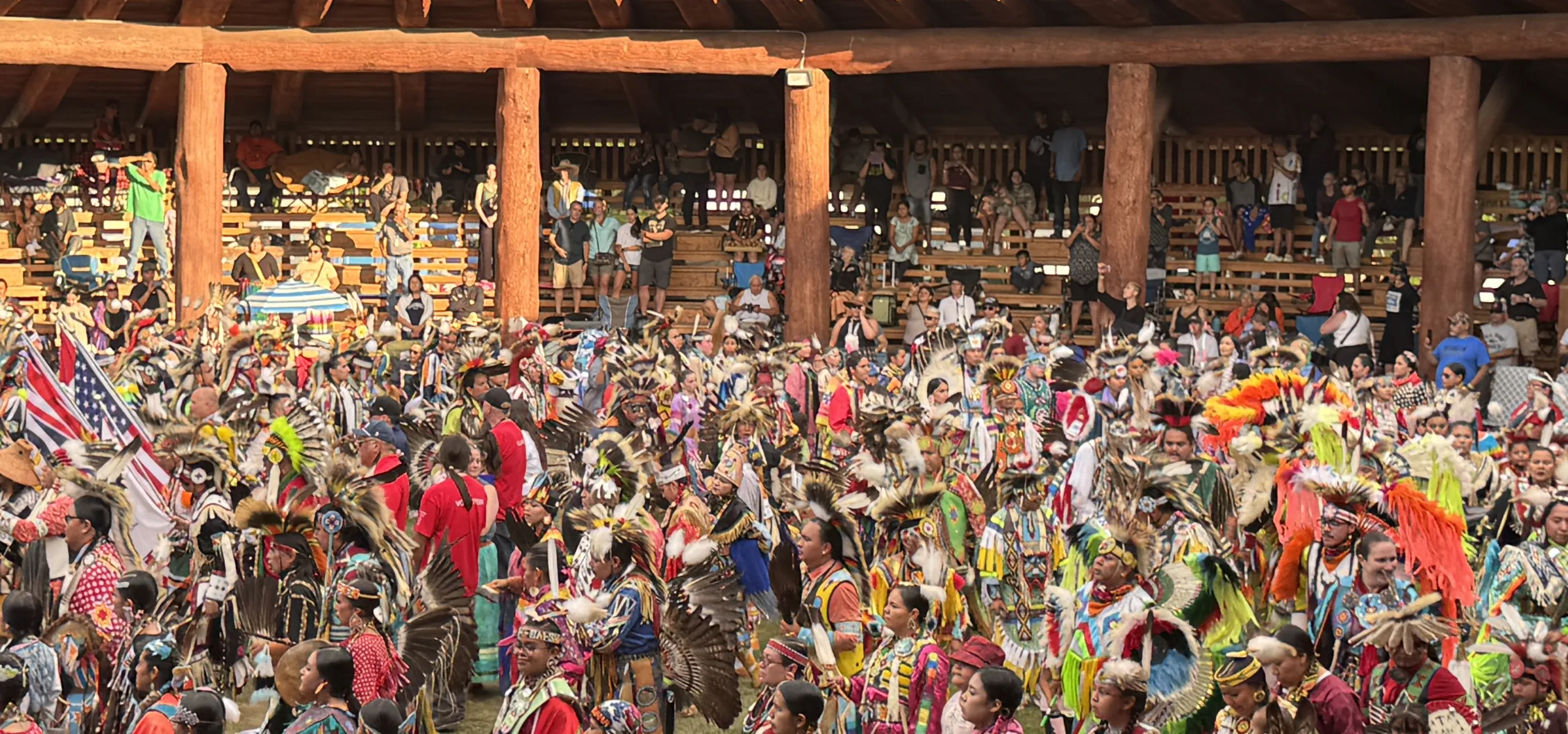Large group of people in colorful traditional regalia participate in a powwow dance inside a wooden arbour, with spectators seated in the background.