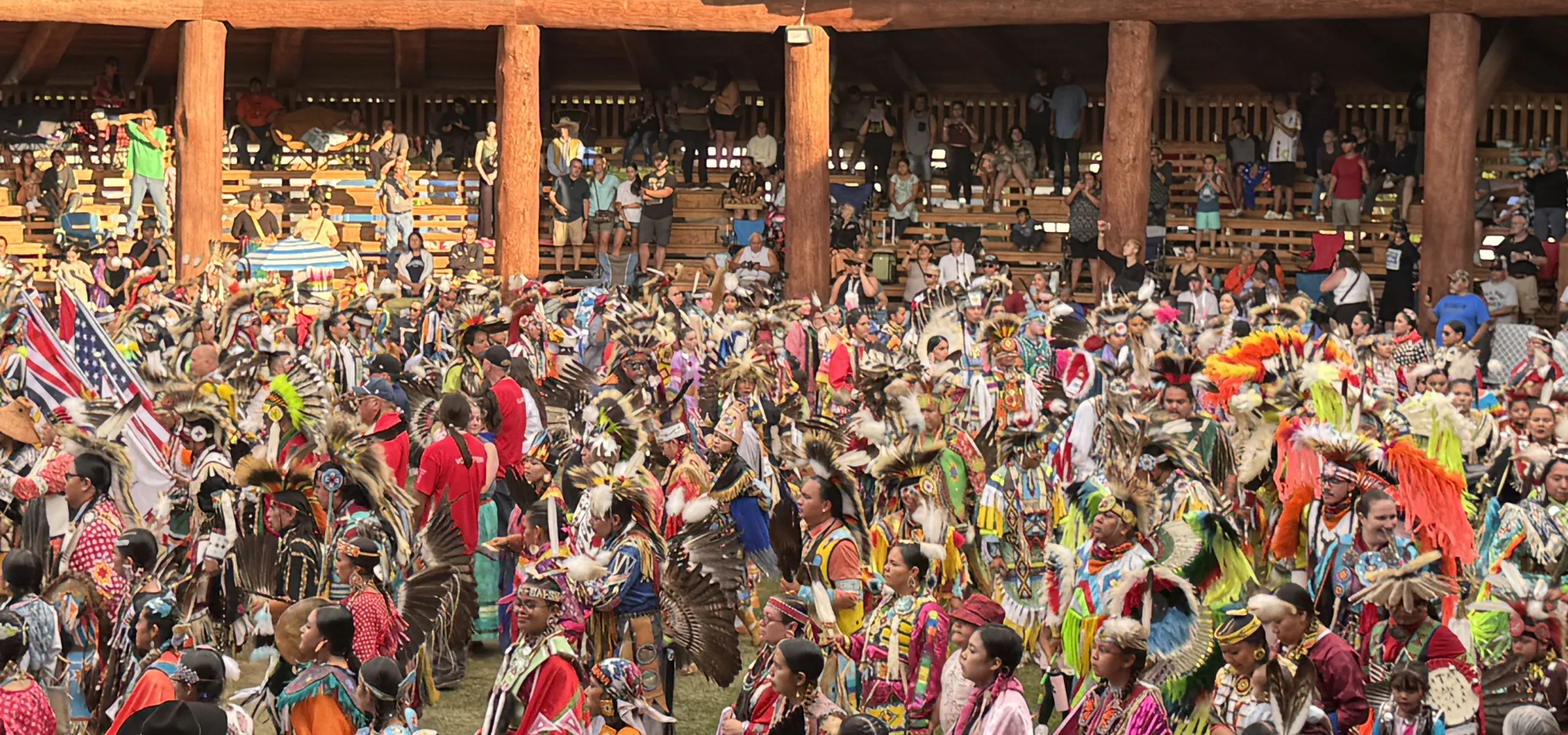 Large group of people in colorful traditional regalia participate in a powwow dance inside a wooden arbour, with spectators seated in the background.