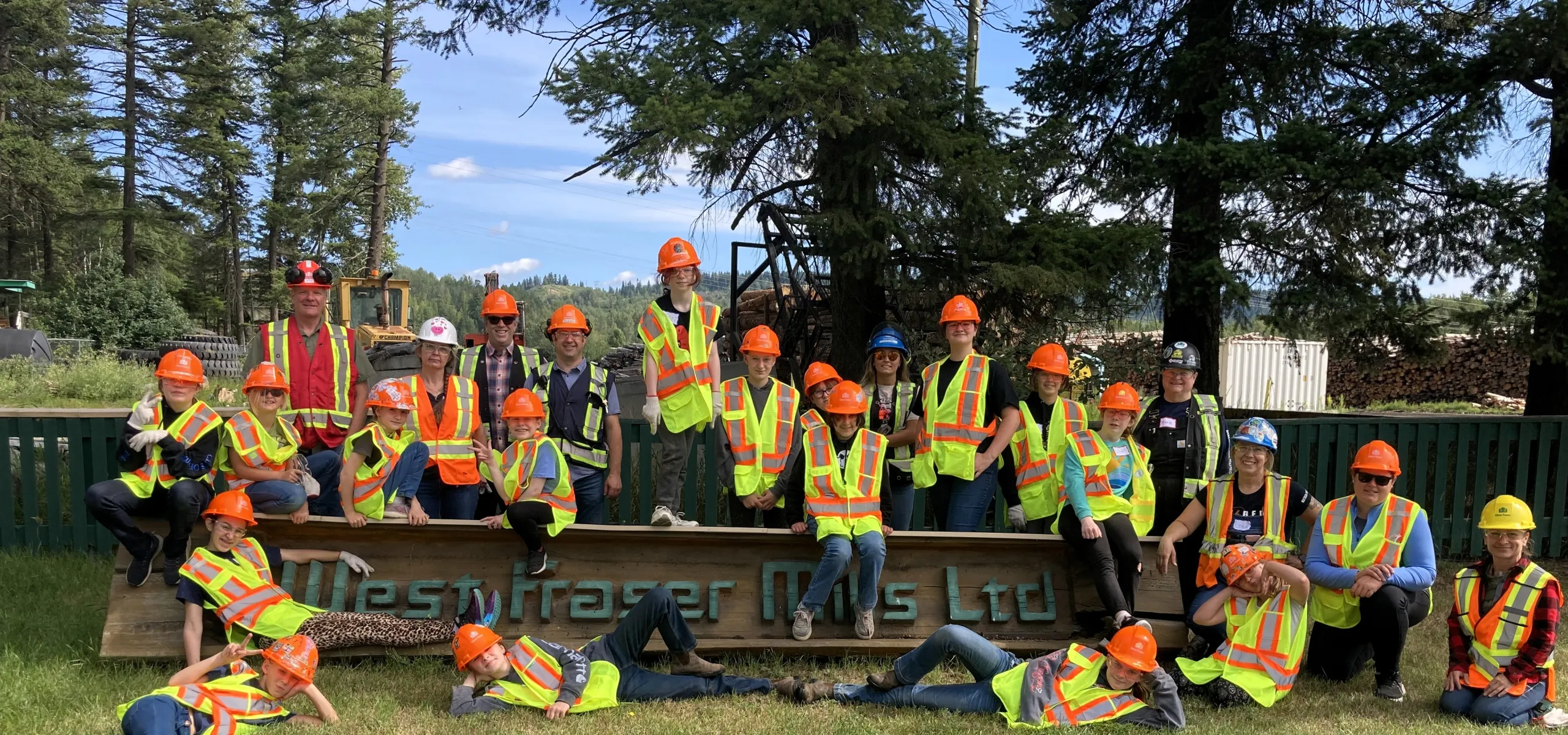 A group of kids wearing safety vests and hard hats pose together outdoors in front of a sign reading "West Fraser Mills Ltd." surrounded by trees.