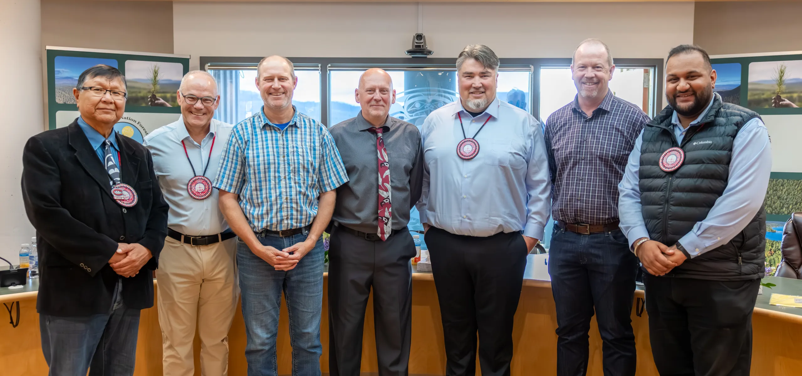 Seven men stand indoors in a row, dressed in business casual and formal attire, with meeting materials visible in the background.
