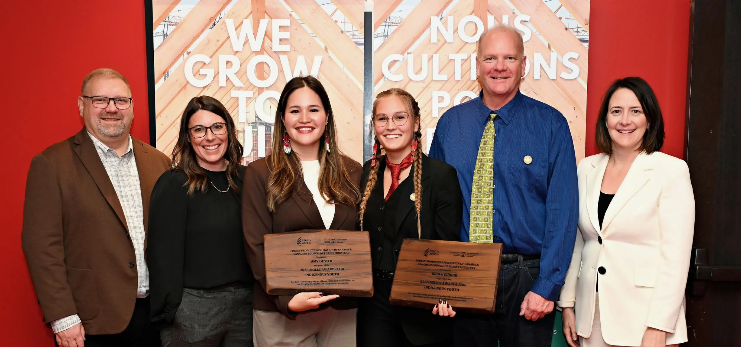 Six people stand indoors, with two women in the center holding award plaques. They are dressed in business attire and are smiling at the camera. Banners and a red wall are in the background.