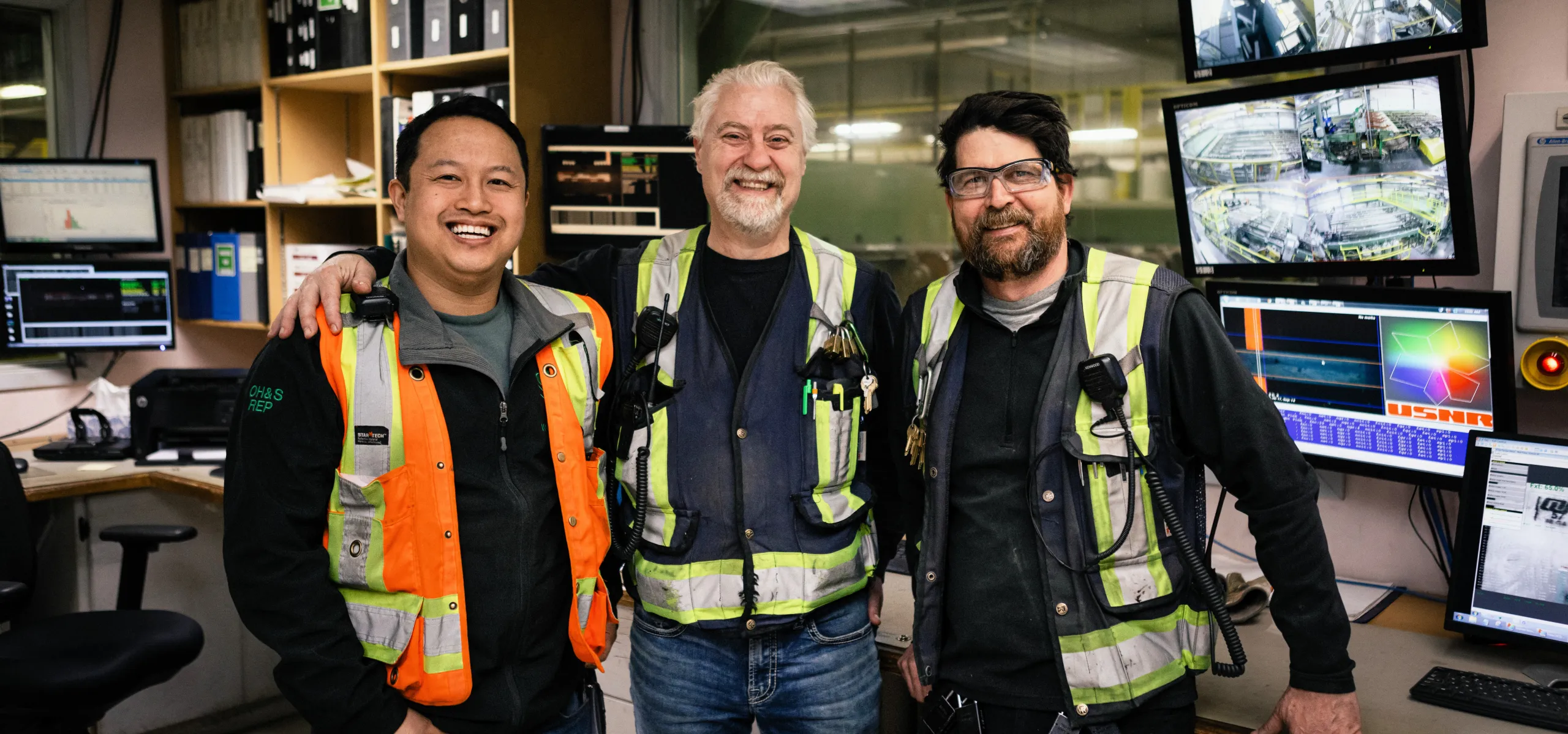 Three men wearing safety vests stand together and smile in a control room with computer monitors and equipment in the background.