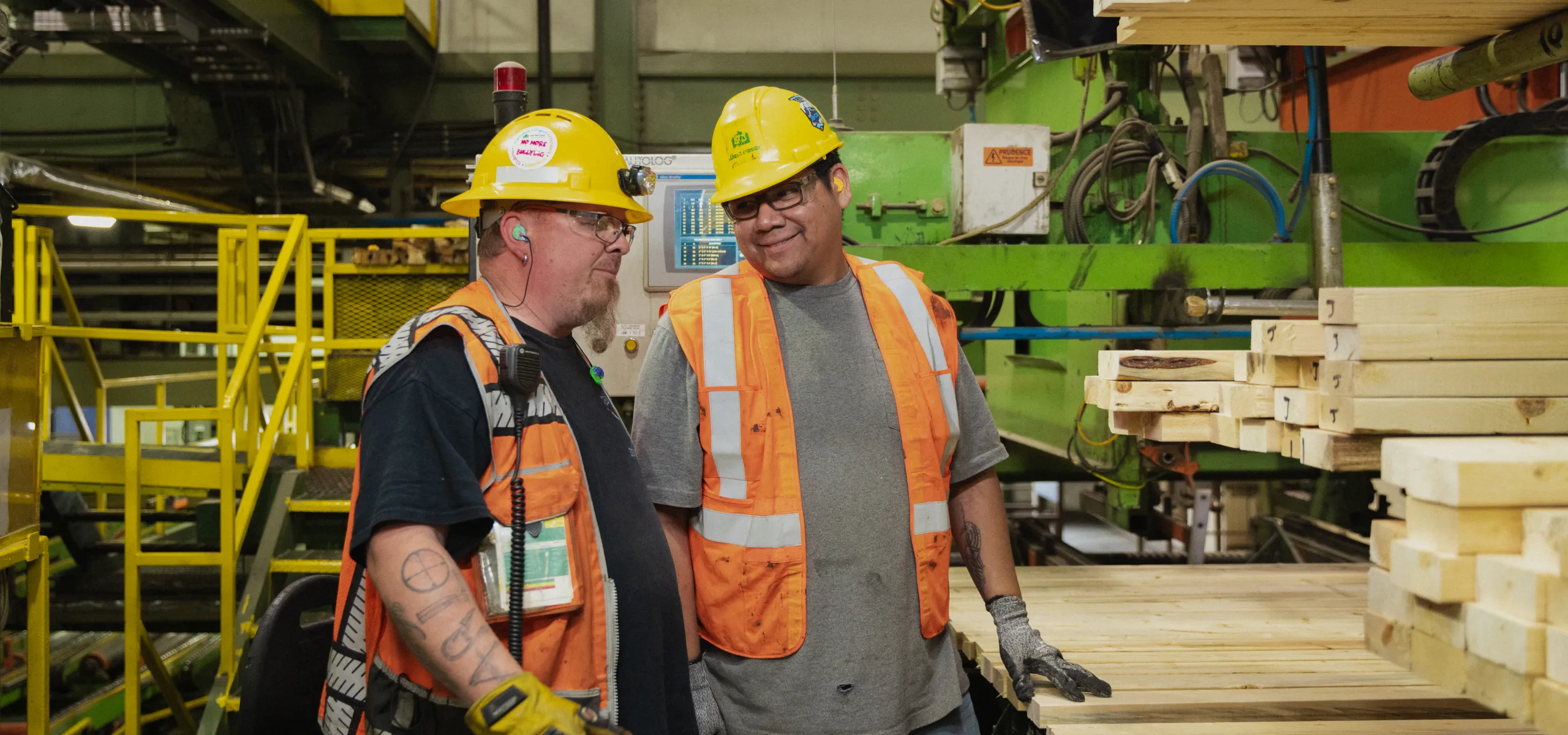 Two construction workers wearing hard hats and safety vests stand beside a stack of wooden planks inside an industrial facility, engaged in conversation.
