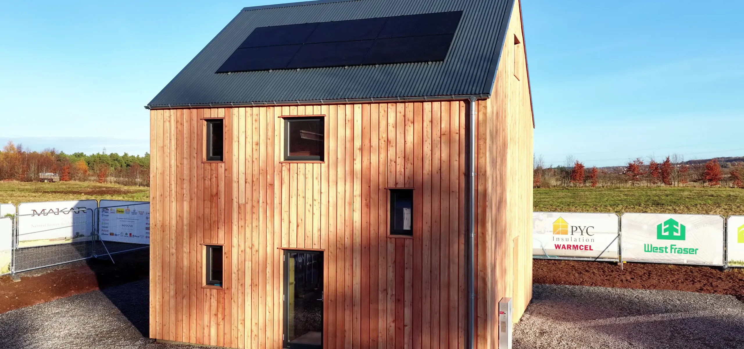 A two-story wooden house with a metal roof and solar panels, surrounded by construction barriers on a gravel lot in a rural area.