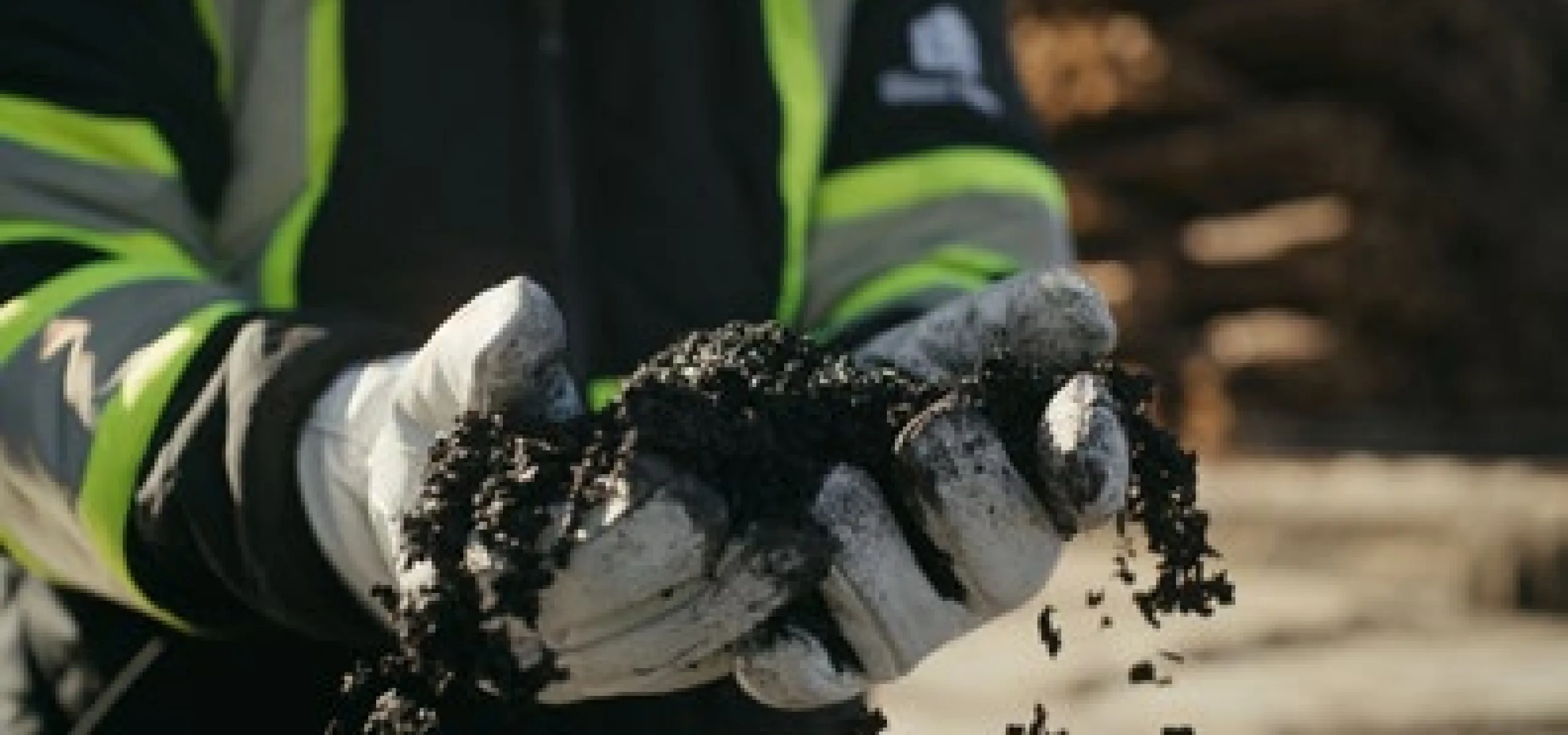 A person wearing gloves and a reflective jacket holds a handful of biochar outdoors, with stacked logs blurred in the background.