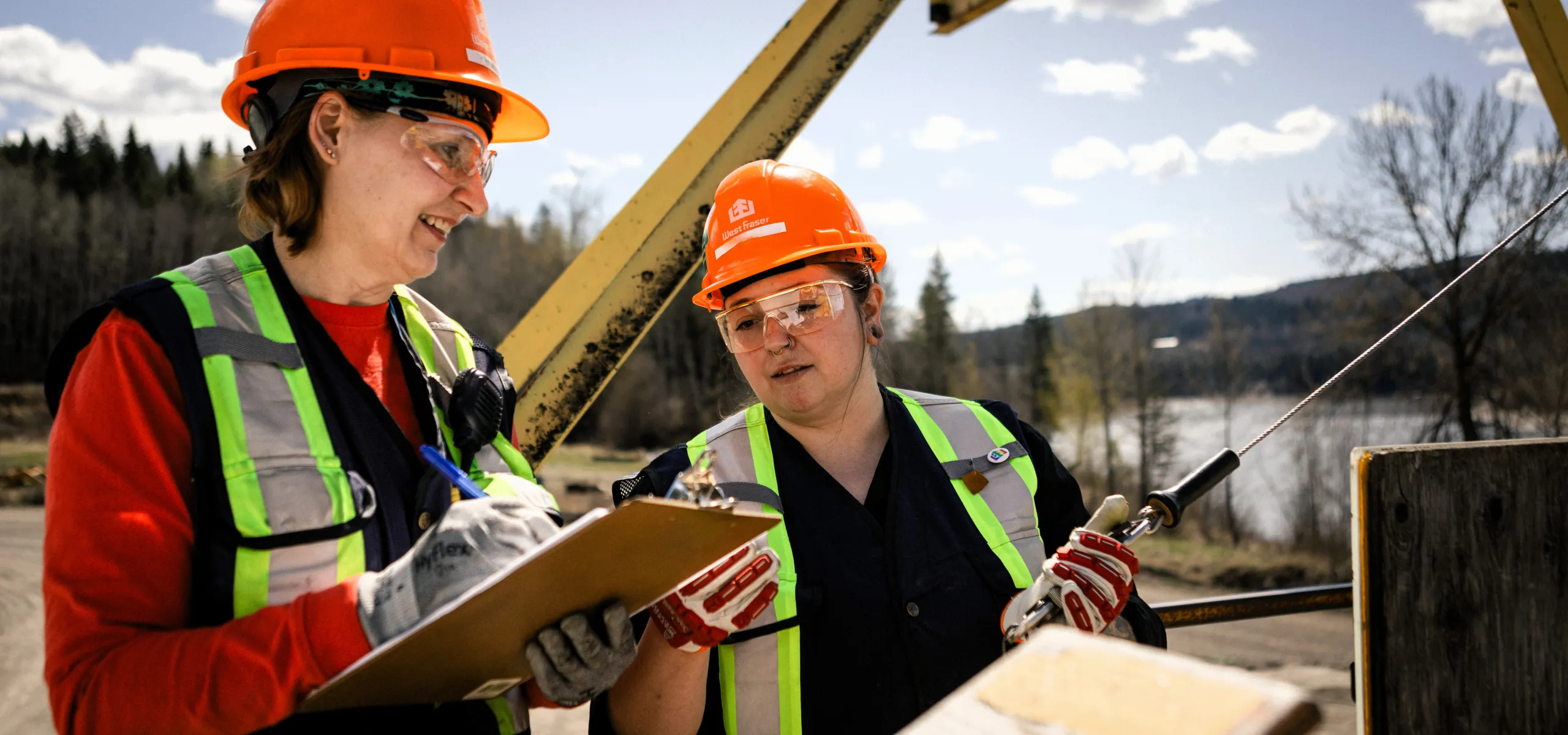 Two workers in safety vests and helmets stand outdoors, one holding a clipboard and pen, the other holding a tool, with trees and water in the background.
