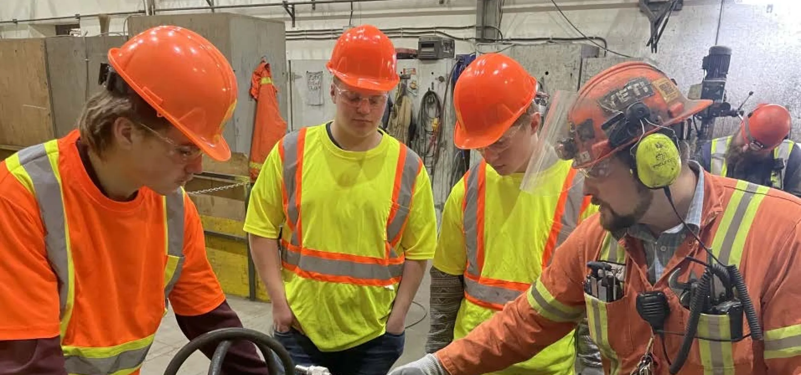Three students and one worker in safety gear examine machinery components on a table in an industrial workshop setting.