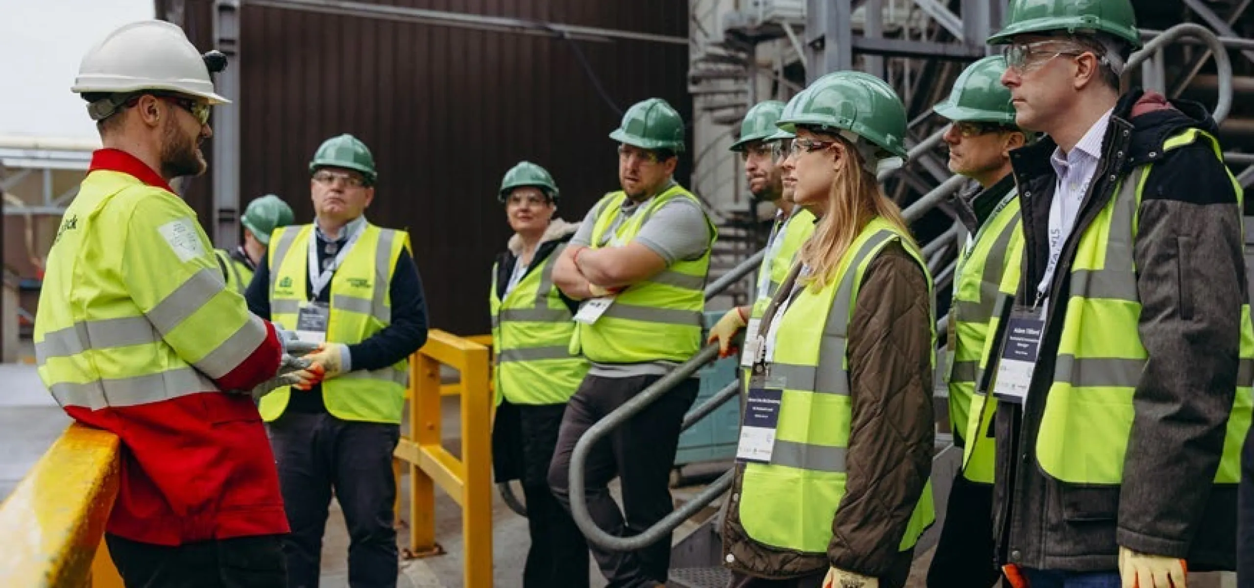 A group of people wearing hard hats, safety vests, and gloves listen to a worker in a red jacket at an industrial site.