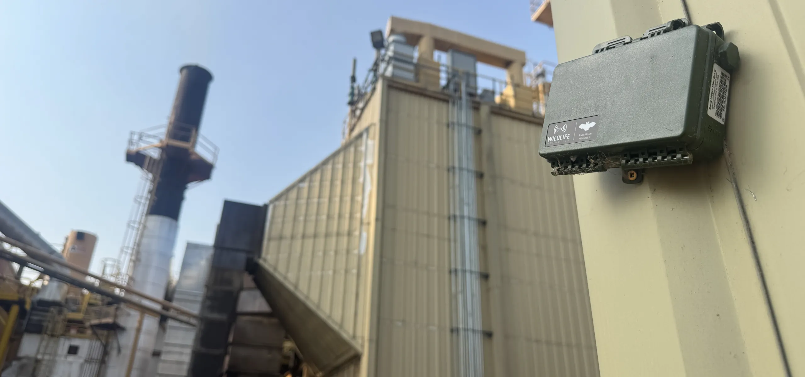 A gray Wildlife Acoustics audio recorder mounted on a metal industrial structure at a facility with smokestacks.