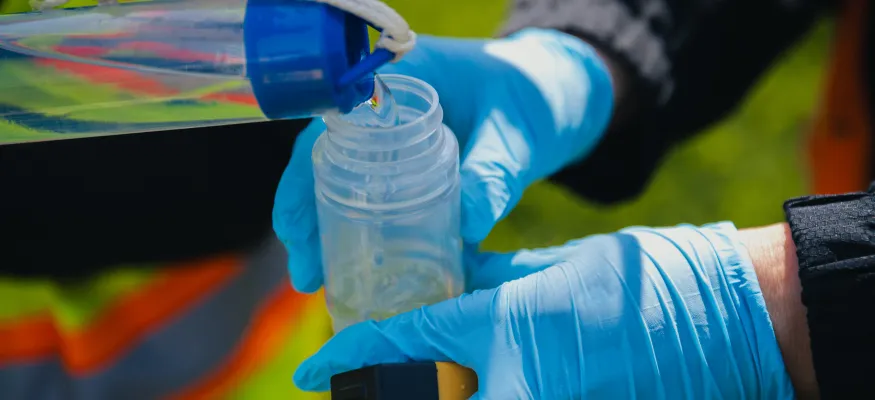 Two gloved hands transfer liquid from a container into a smaller bottle outdoors.